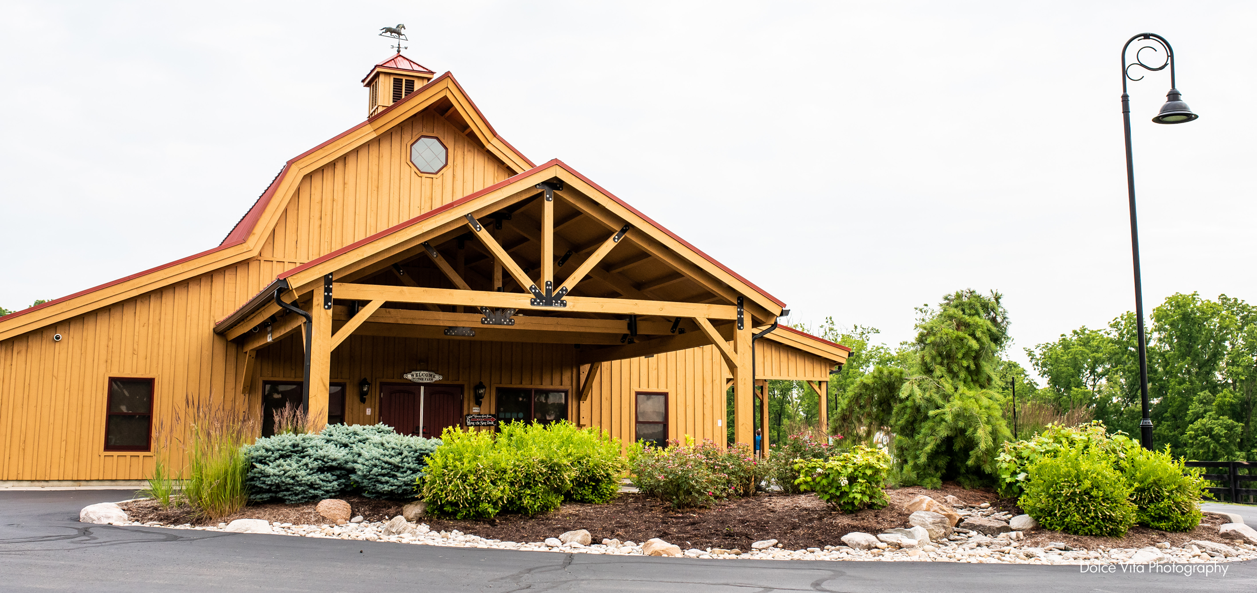 A wooden building with a red roof and a weather vane on top, surrounded by greenery and a landscaped garden with rocks and bushes.