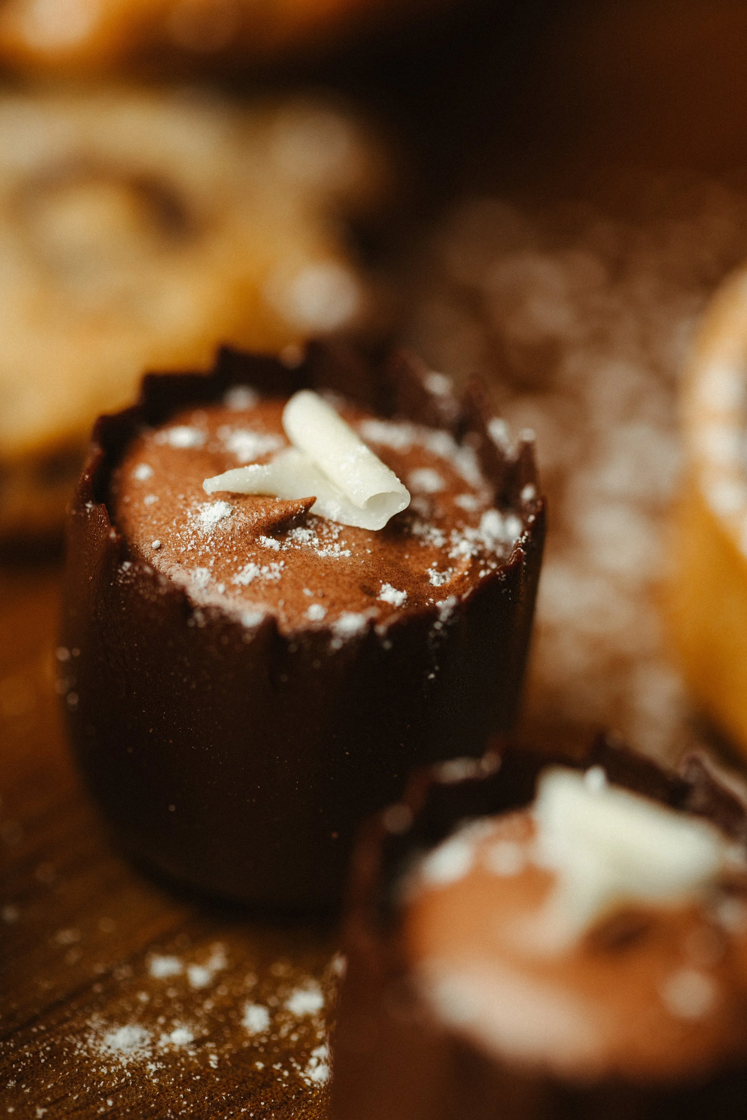 Close-up of a chocolate dessert garnished with white chocolate shavings and powdered sugar, with a blurred background.