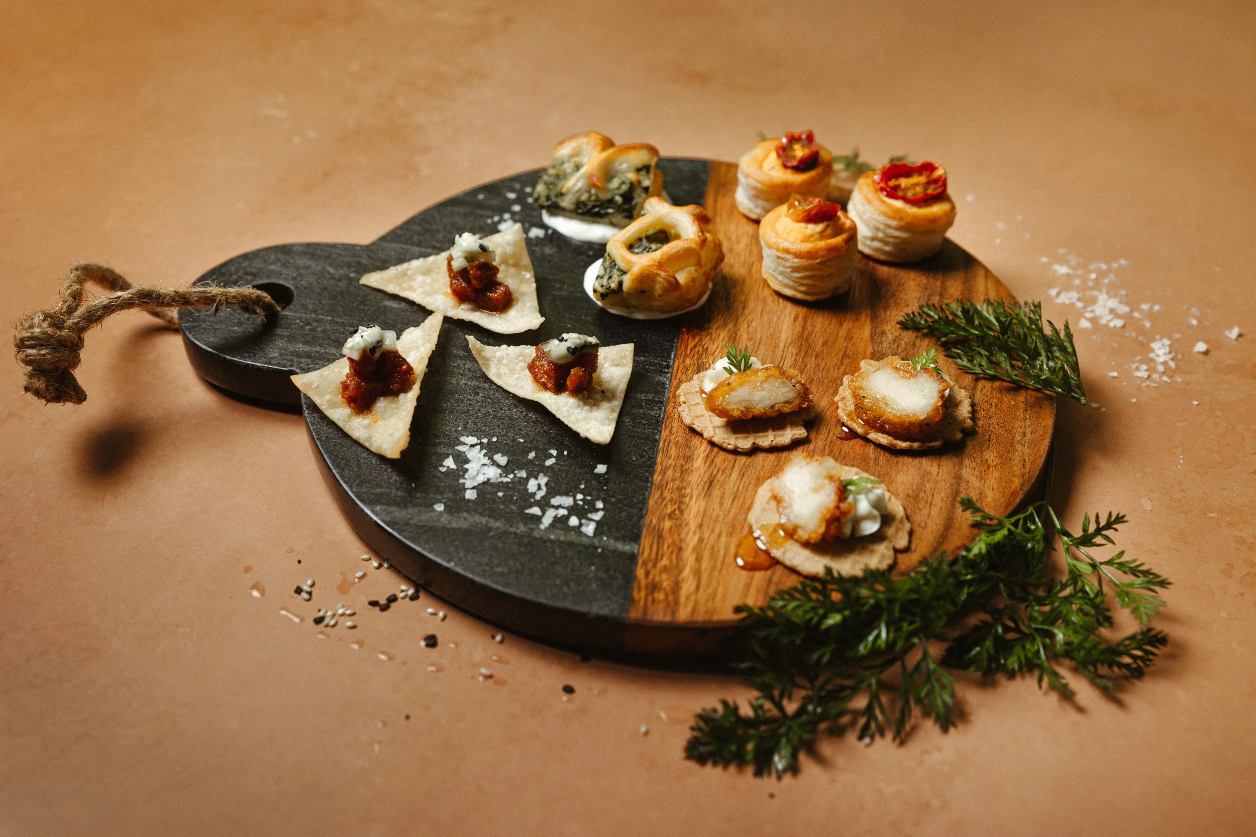 Assorted small appetizers on a black and wooden serving tray with scattered salt and herbs.