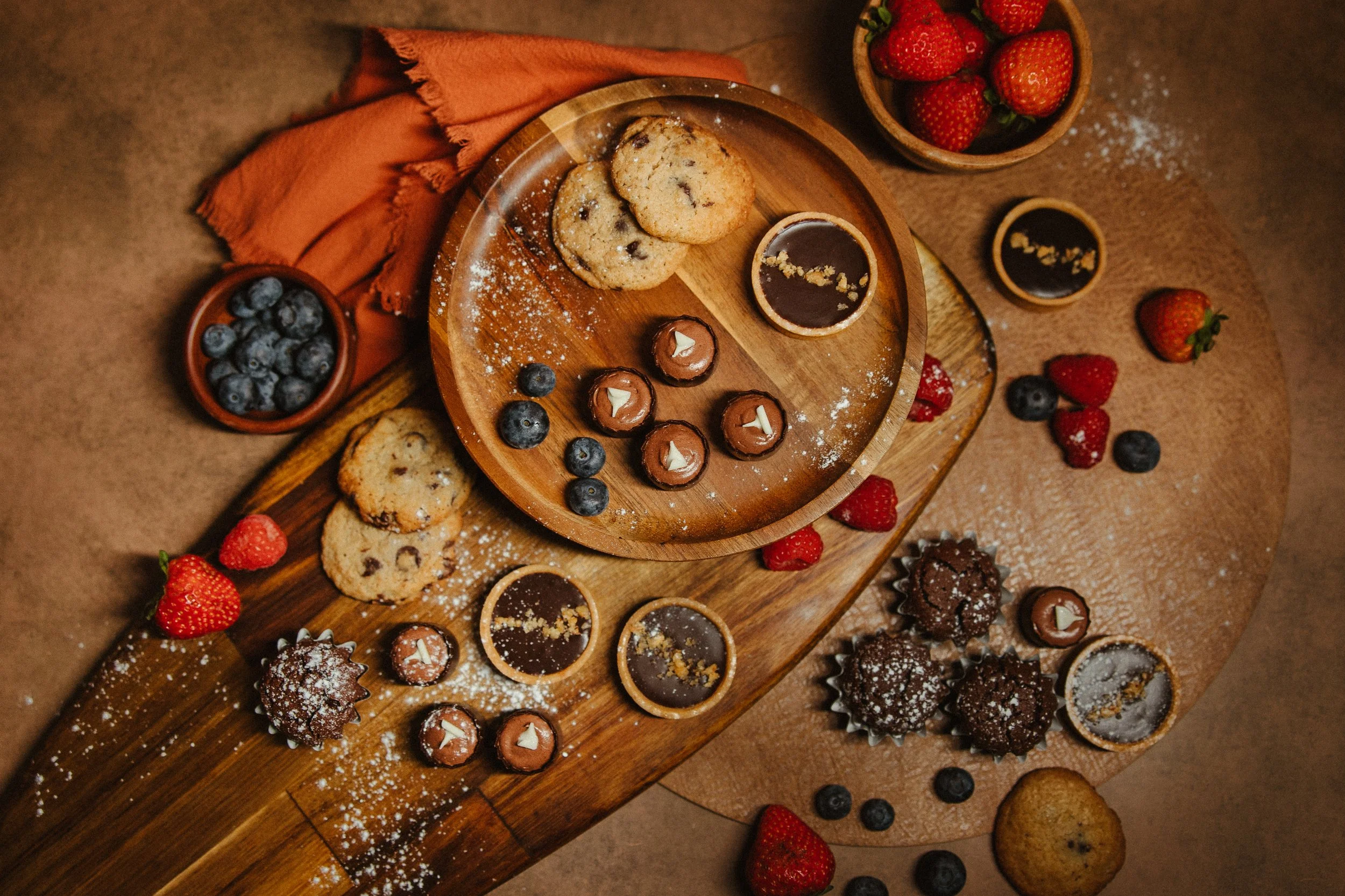 Assorted chocolate chip cookies, strawberries, blueberries, and small bowls of chocolate and berry desserts on a wooden platter and table.