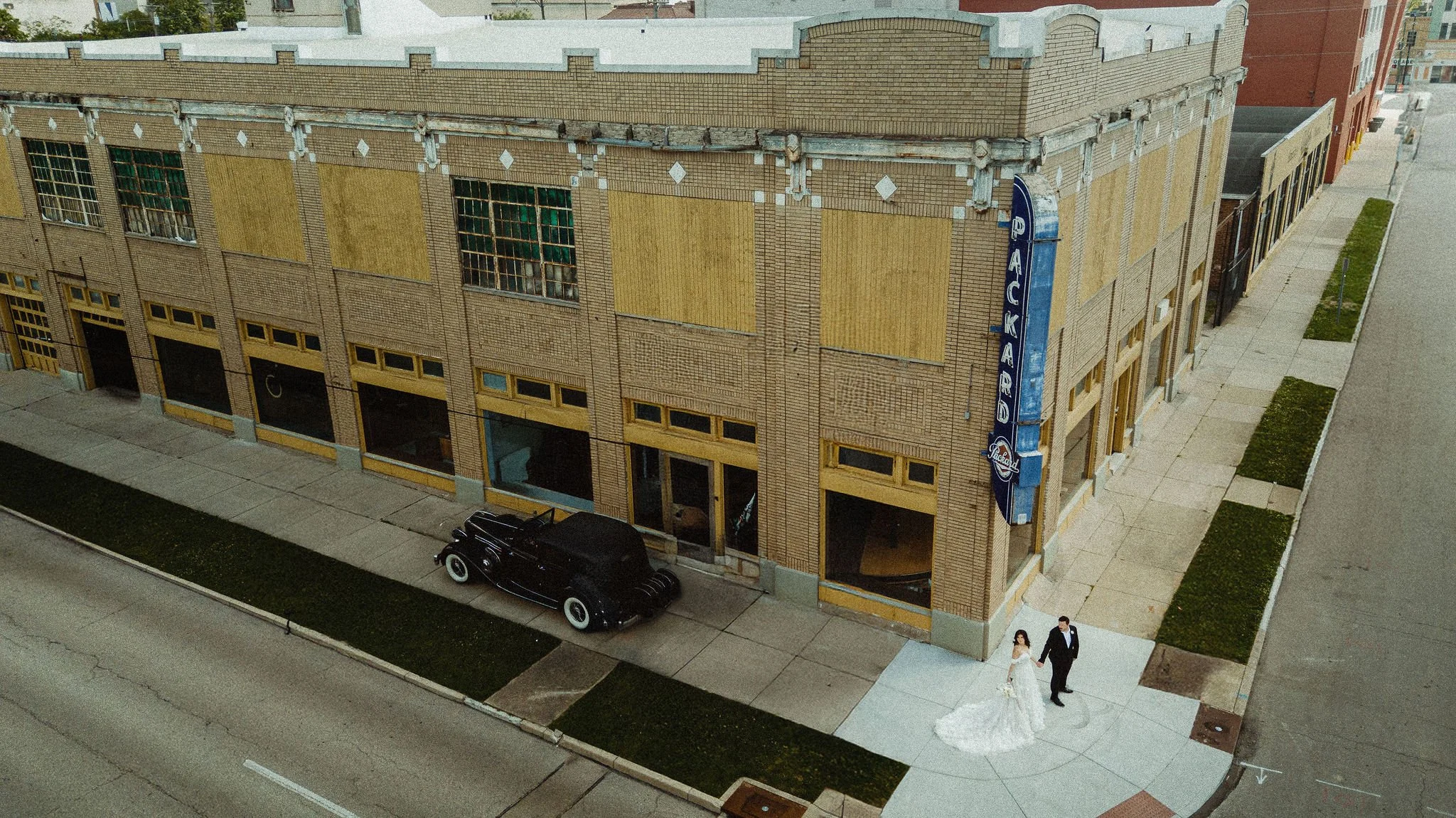 A vintage black car parked on the street in front of a brick building with yellow accents, large windows, and a blue sign that reads 'Packard' on the side.