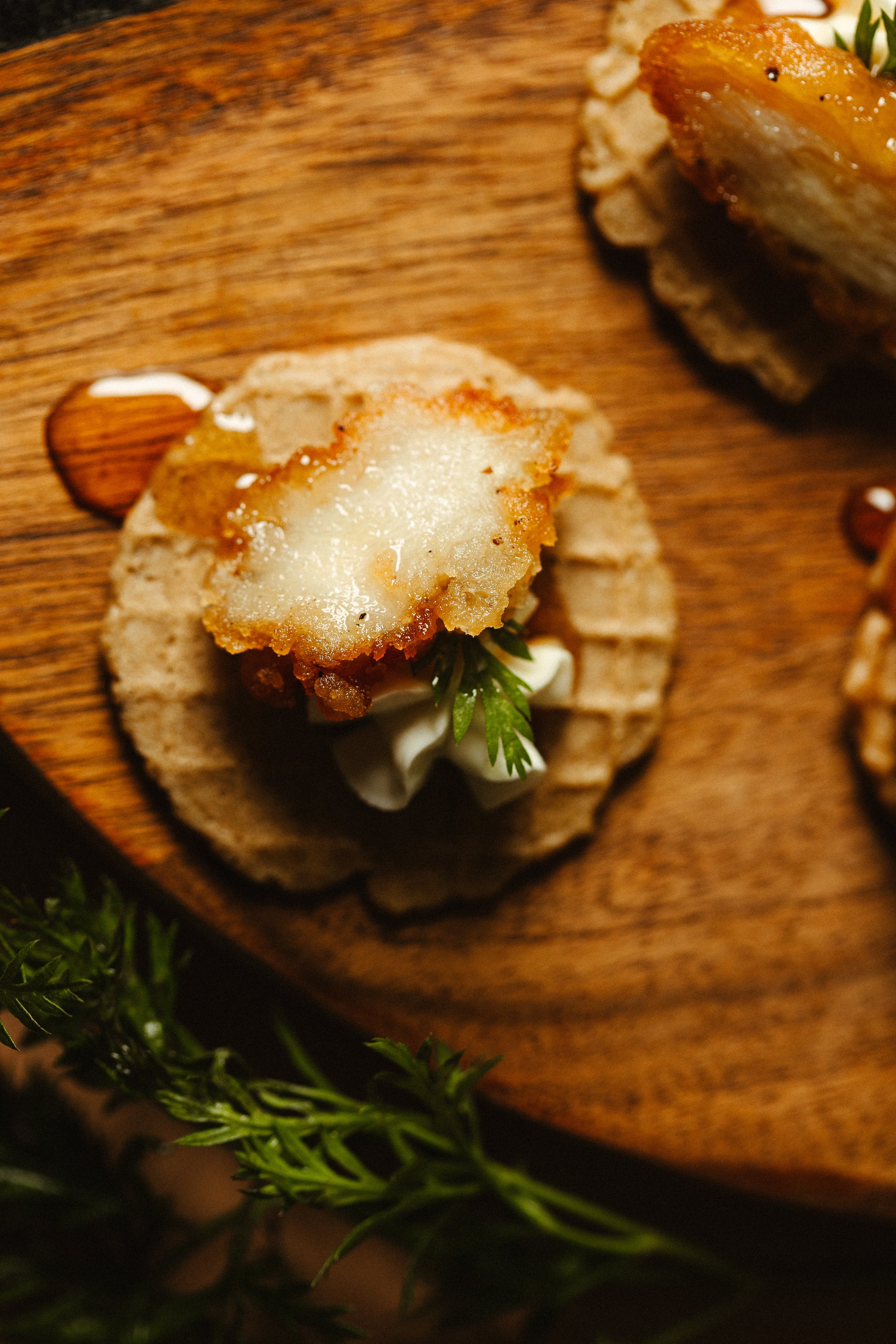 A close-up of an appetizer featuring a crispy fried piece of food topped with a light sauce, placed on a round bread or cracker on a wooden serving board, with some greenery in the background.