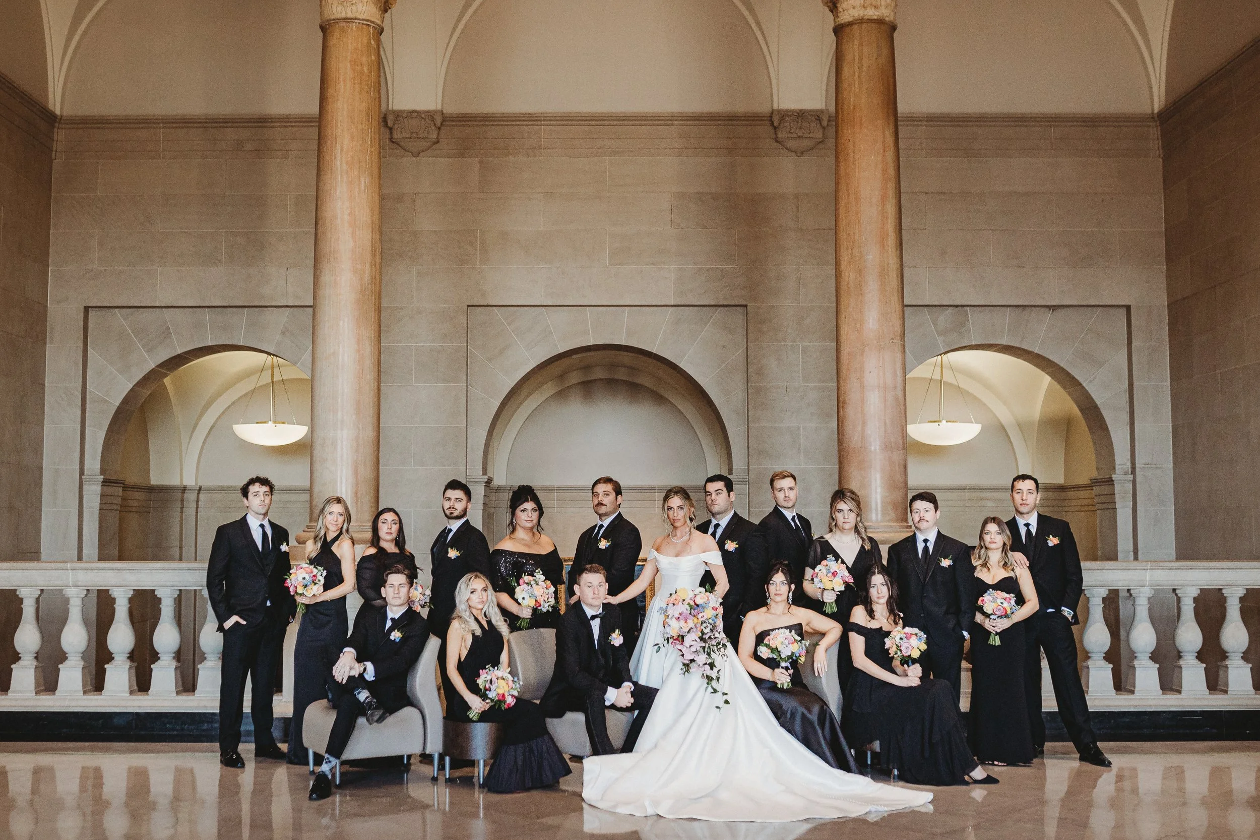 A large group of people, including men and women, dressed in formal attire, posing for a wedding photograph on a grand staircase inside a historic building with high ceilings, large columns, and arched doorways.