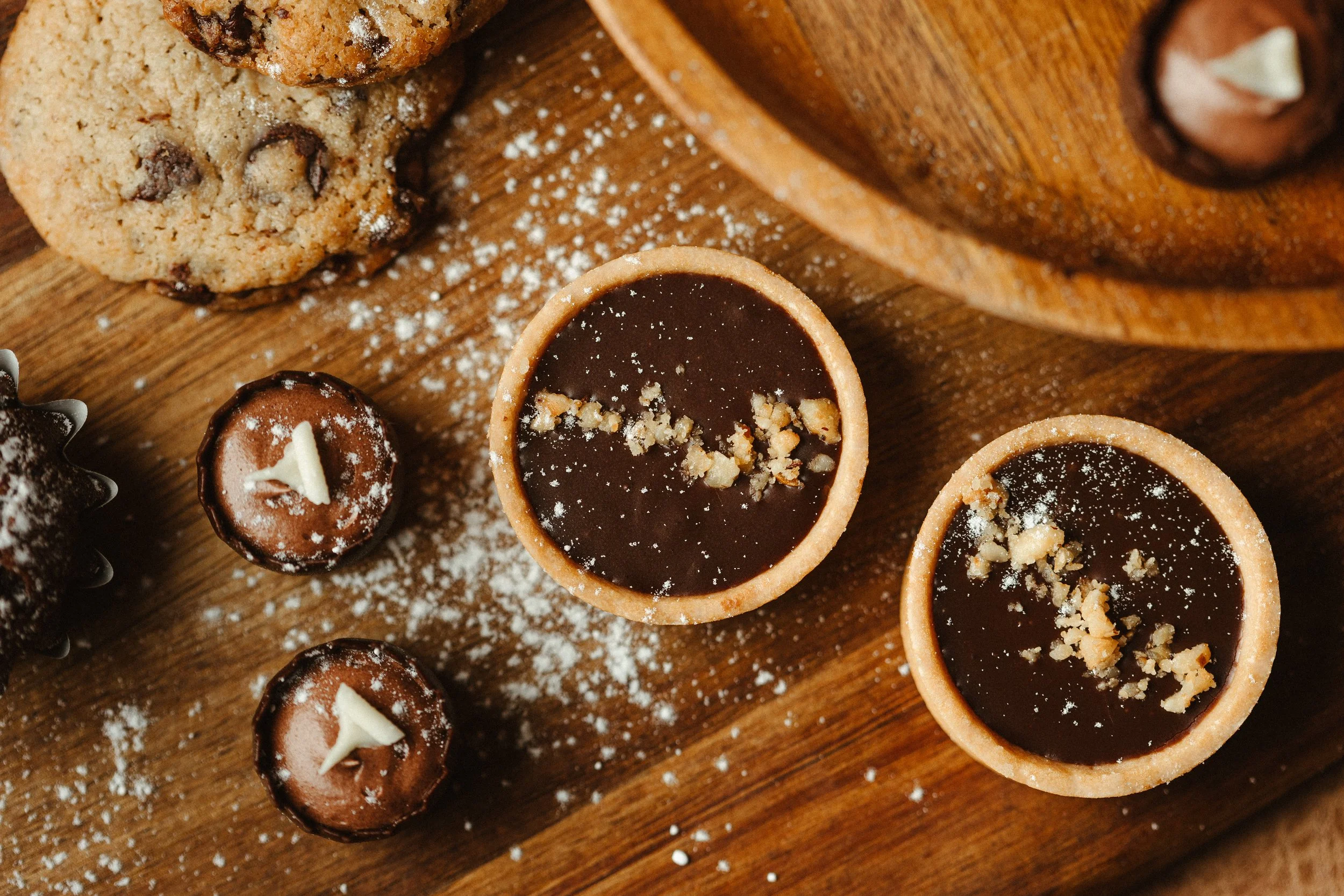 Assorted cookies and chocolate tartlets on a wooden surface, dusted with powdered sugar.