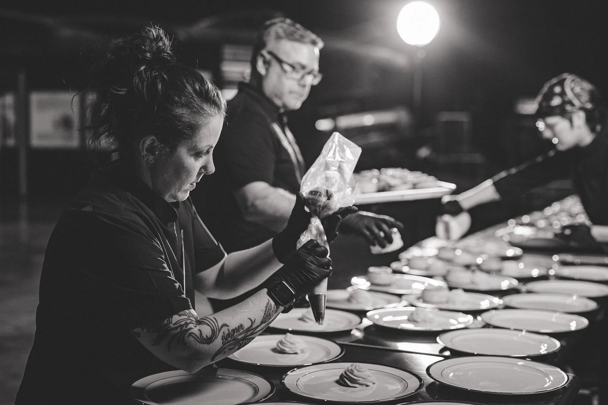 Black and white photo of three people preparing plates on a counter in a dimly lit setting.