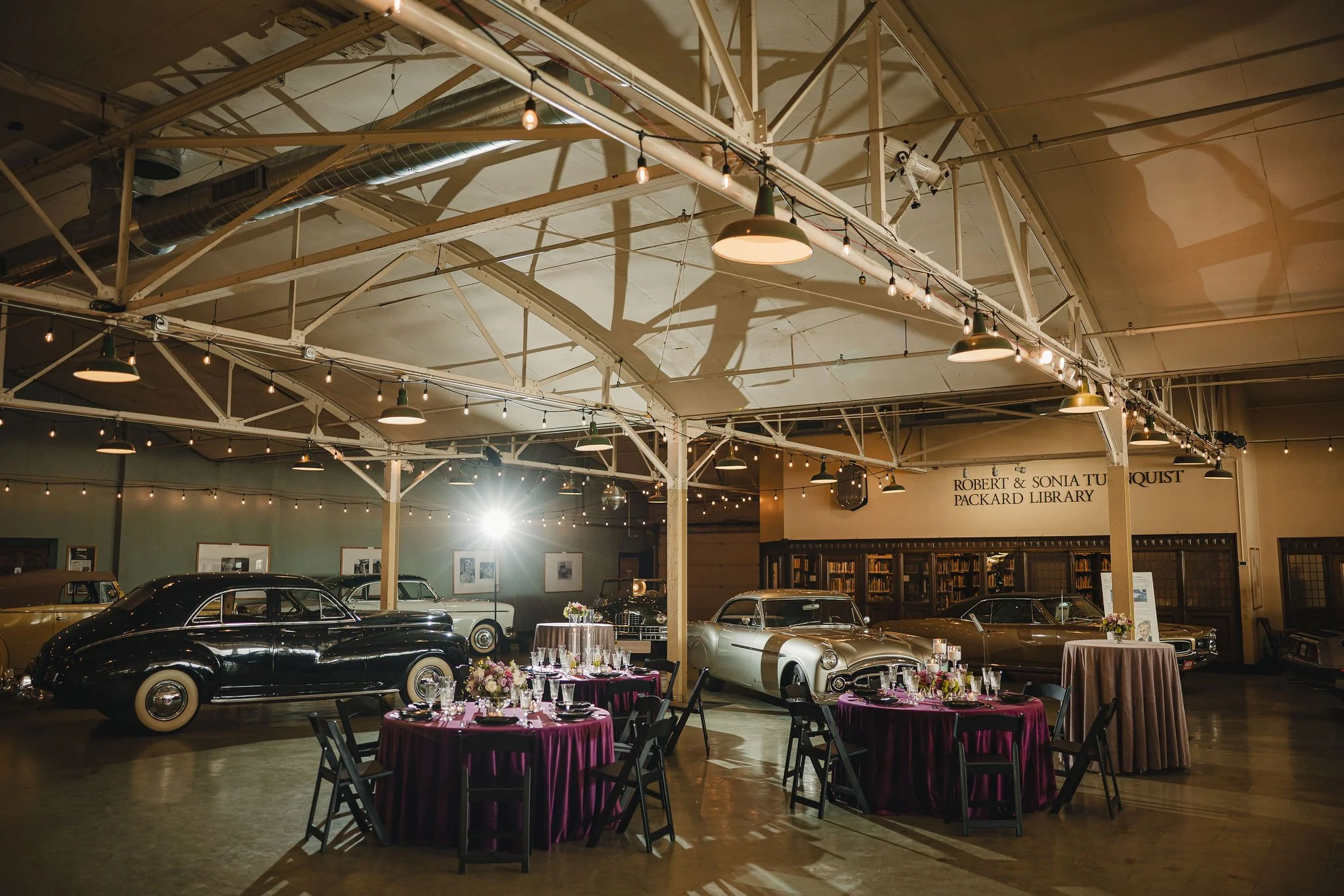 Vintage cars displayed in an indoor space with industrial ceiling, string lights, and tables set for an event with flowers and purple tablecloths.