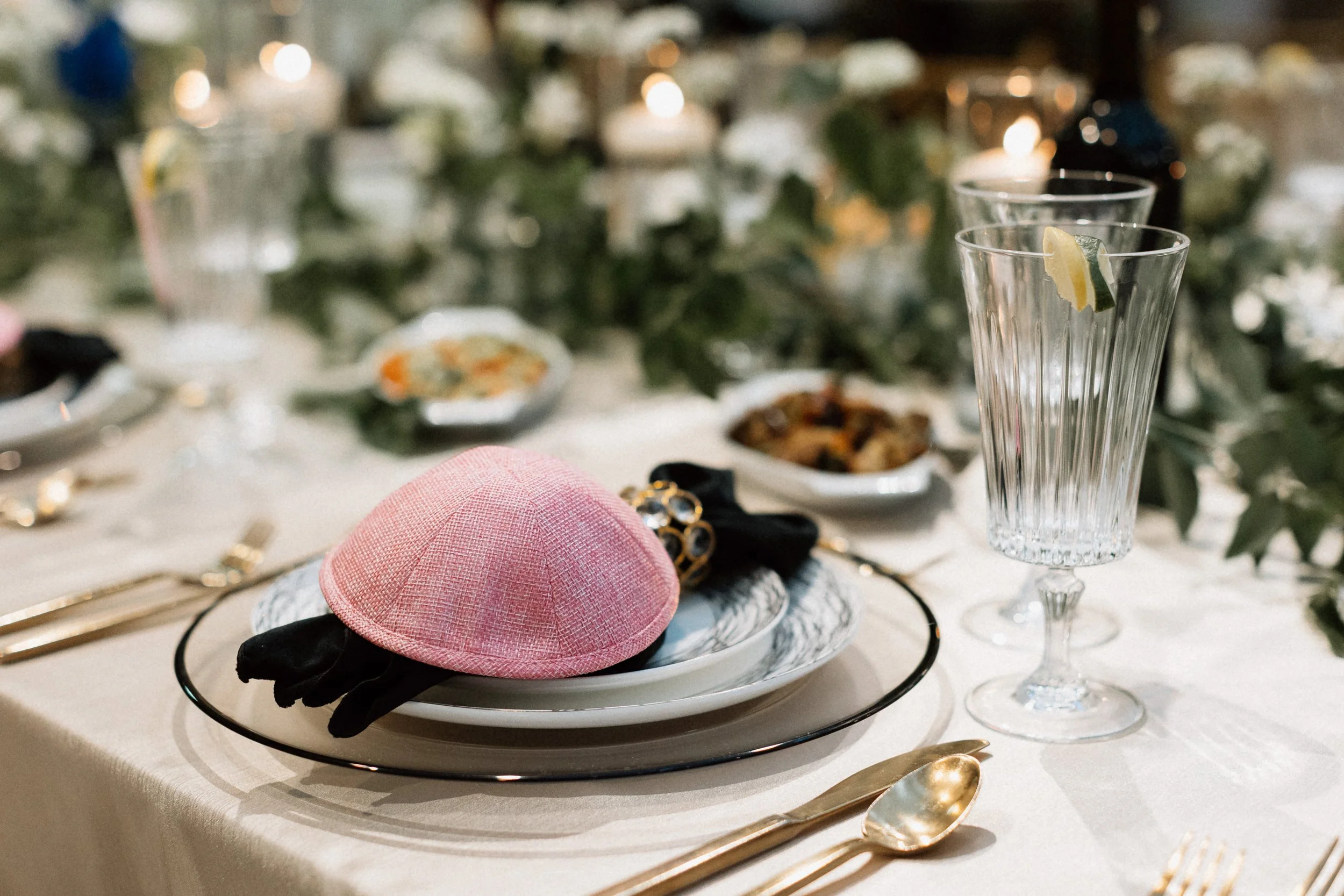 A dining table setup with a glass of lemon water, a pink hat with black ribbon, plates, gold utensils, and blurred food dishes in the background, decorated with greenery and warm lighting.