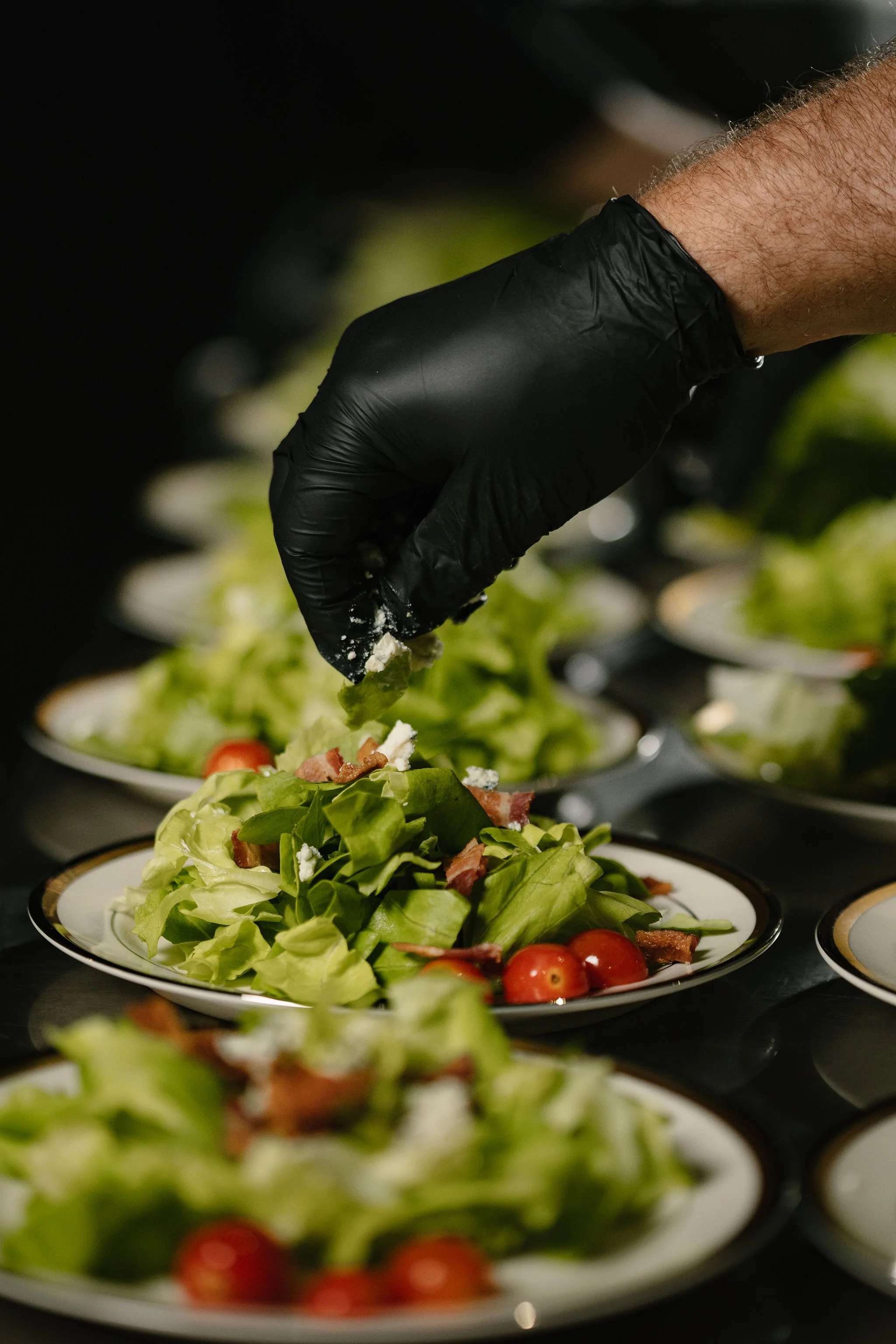 A person wearing a black glove preparing a fresh salad with lettuce, cherry tomatoes, crumbled cheese, and bacon bits on a white plate.
