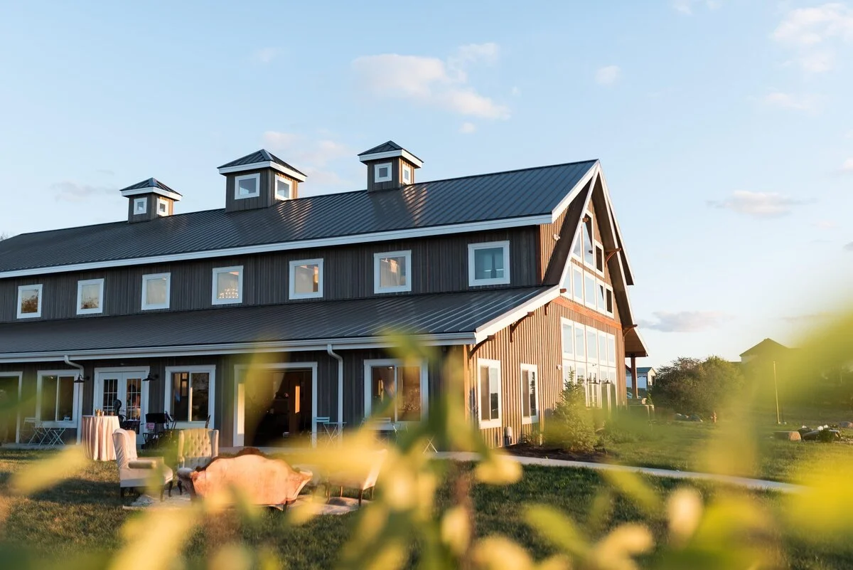 A large modern barn-style house with metal roof and multiple large windows, surrounded by a grassy yard with outdoor furniture, under a partly cloudy sky.