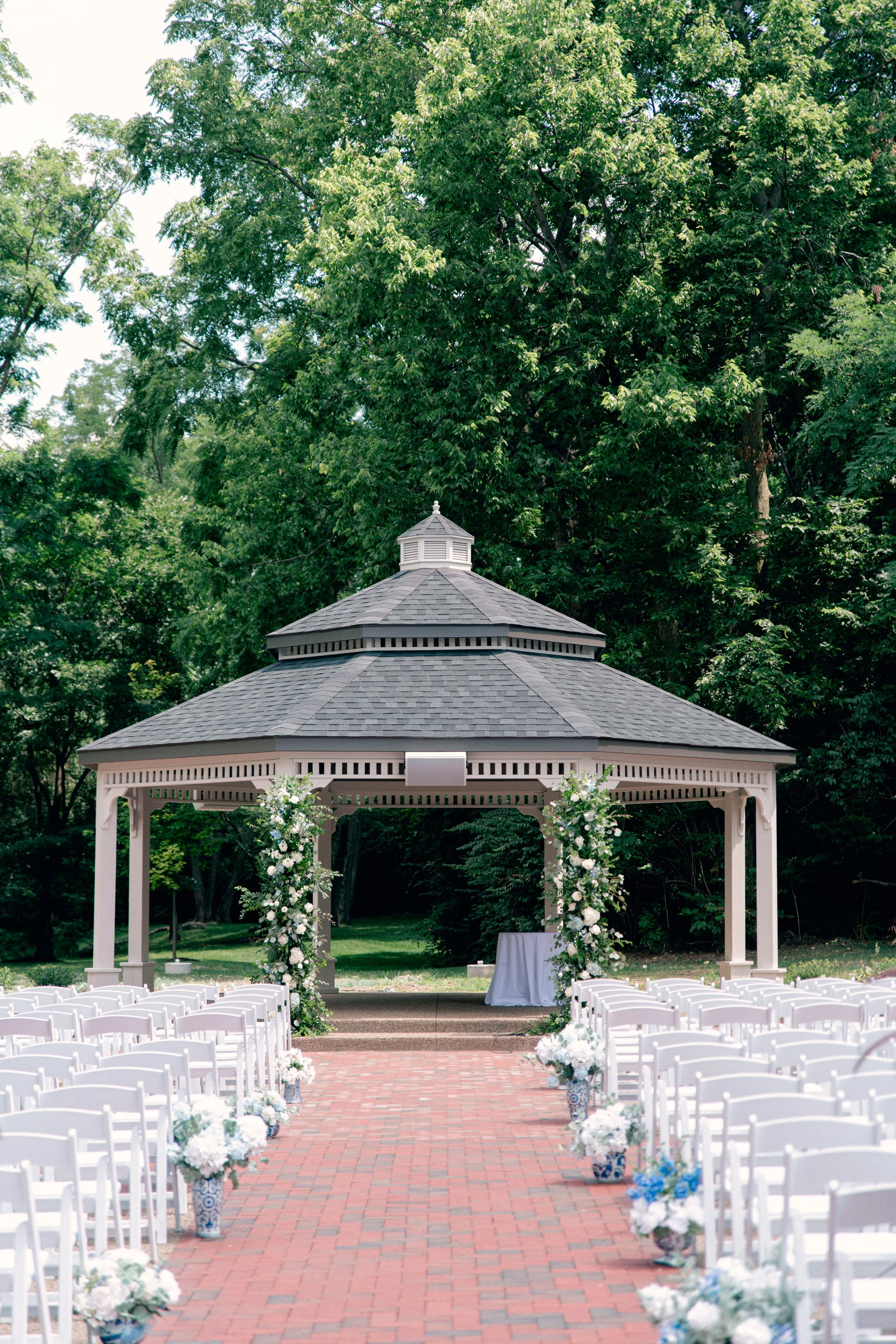 Outdoor wedding ceremony setup with rows of white chairs, decorated with white and blue floral arrangements, facing a gazebo with white pillars on a brick pathway, surrounded by green trees.