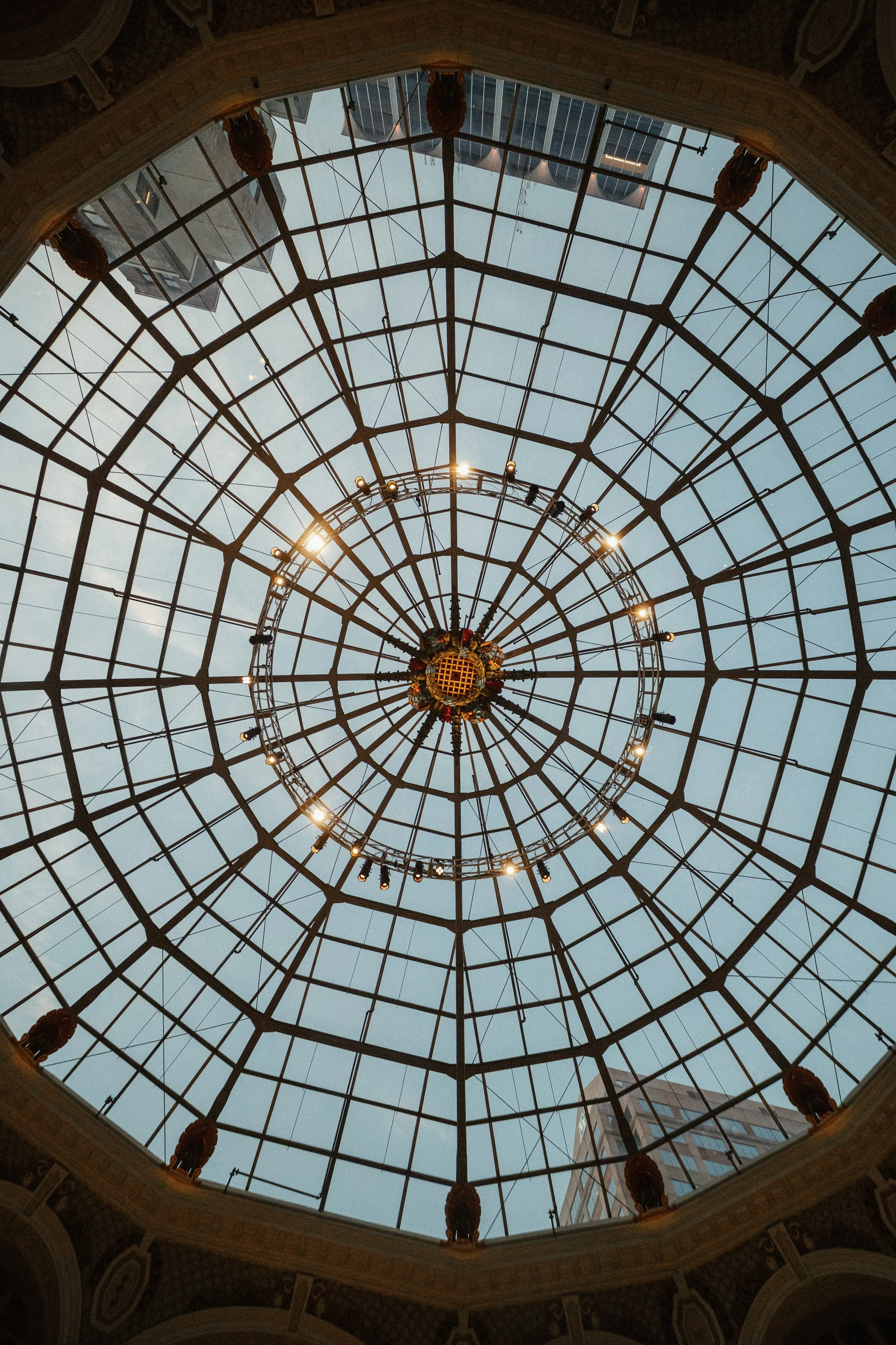 View of a glass-domed ceiling with a circular chandelier and surrounding architectural details, seen from below inside a building.