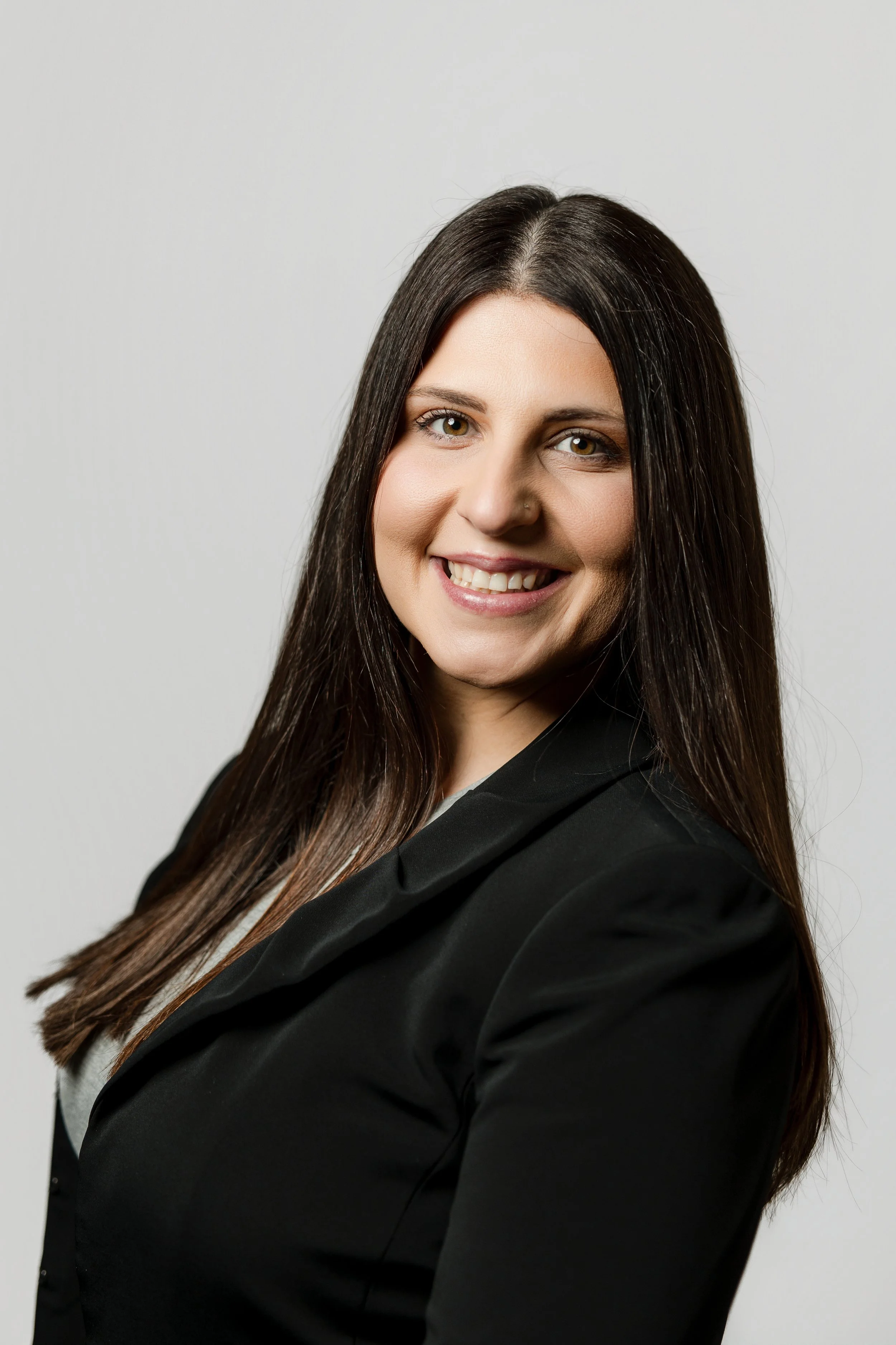 A smiling woman with long dark hair, wearing a black blazer, posing against a plain light gray background.