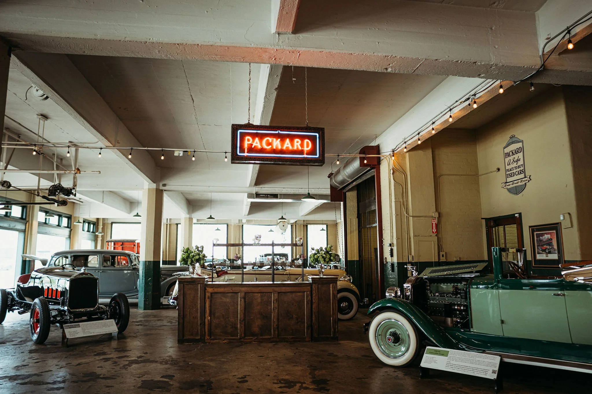 Interior of a vintage car museum with classic cars on display, including a black open-top car and a green car, and a neon sign reading 'Packard' hanging from the ceiling.
