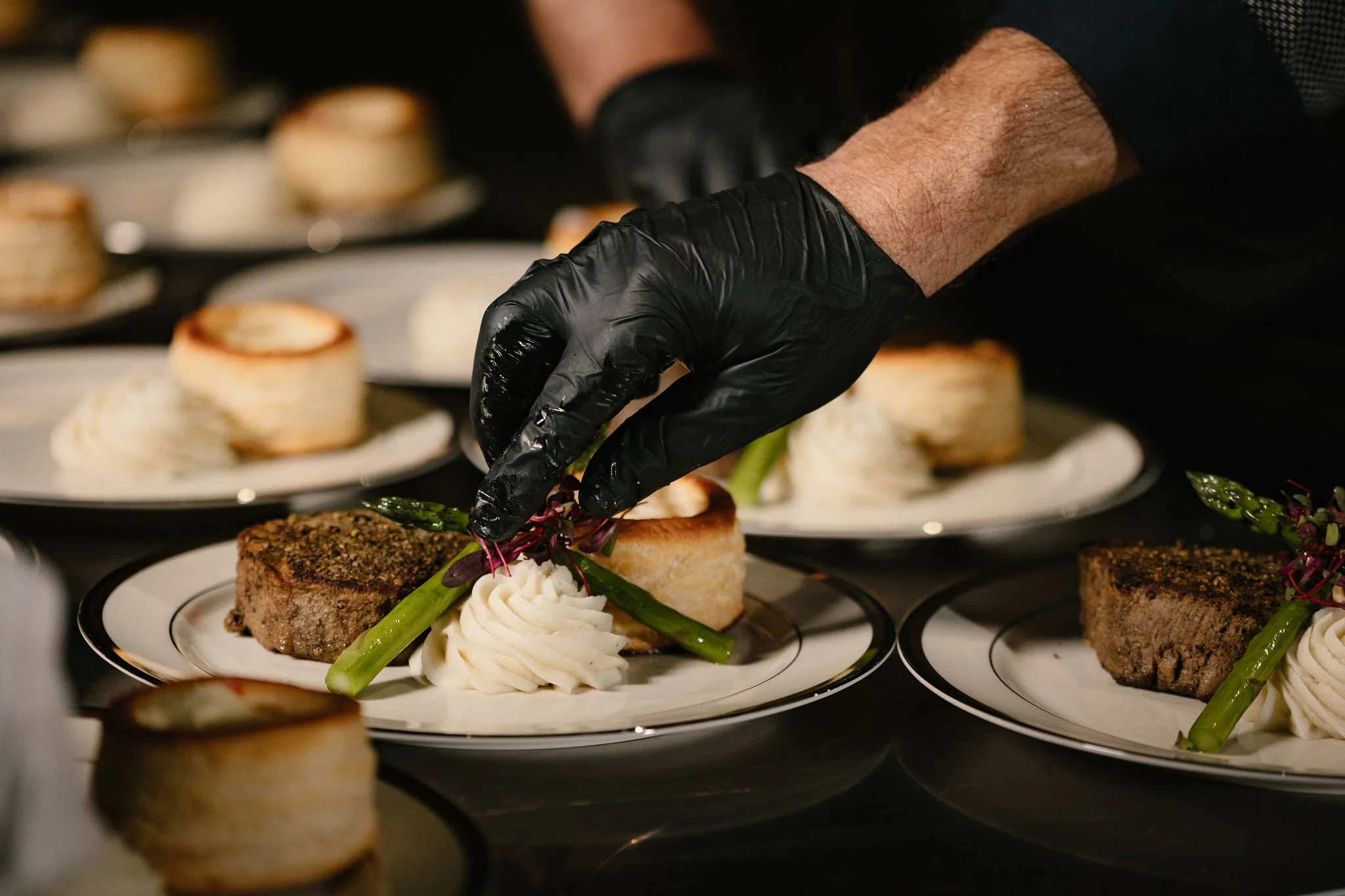 A chef wearing black gloves garnishing plates with food, including a portion of whipped cream, green vegetables, and a meat dish.