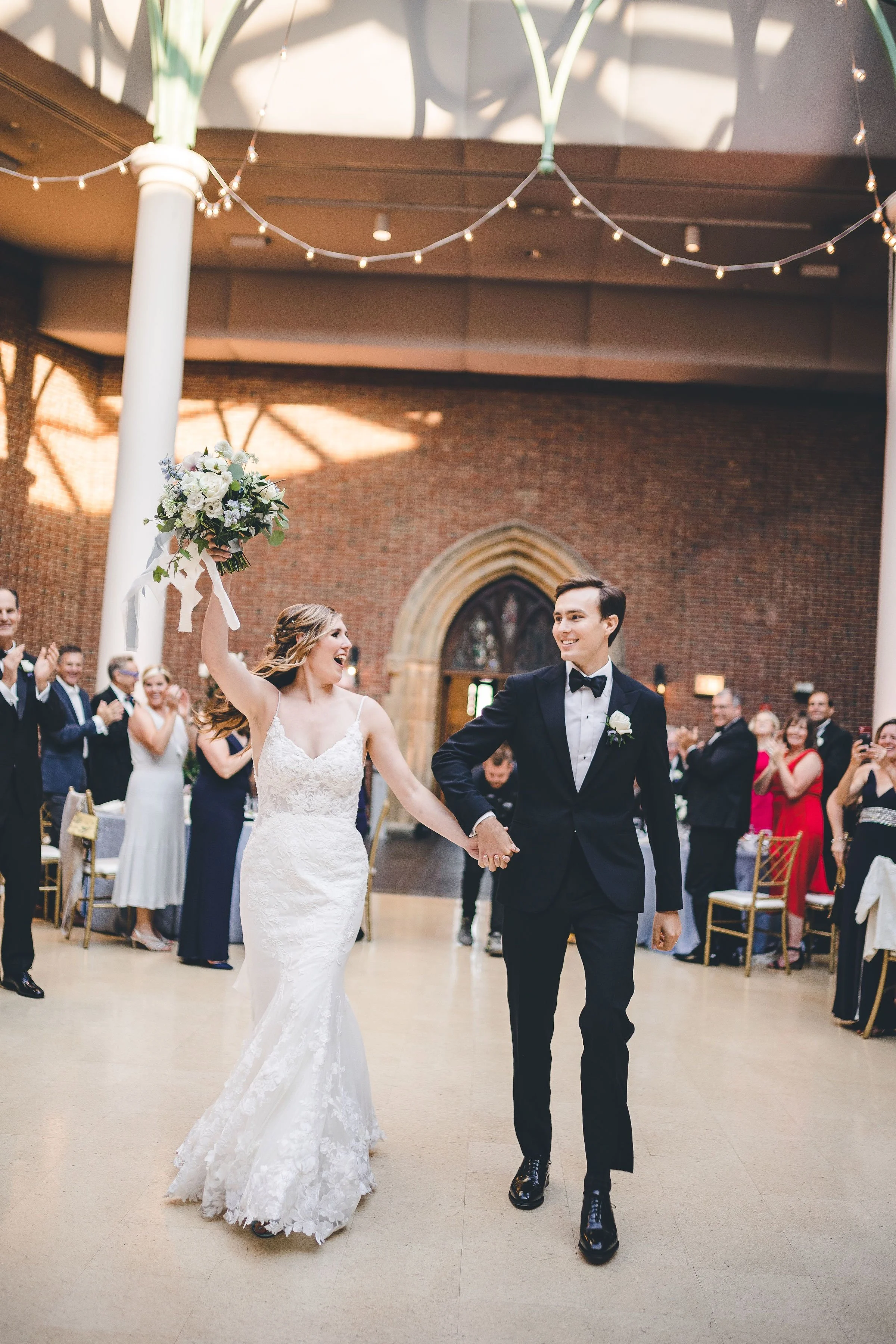 Bride and groom dancing at their wedding reception, surrounded by guests in an indoor hall decorated with string lights and a brick wall