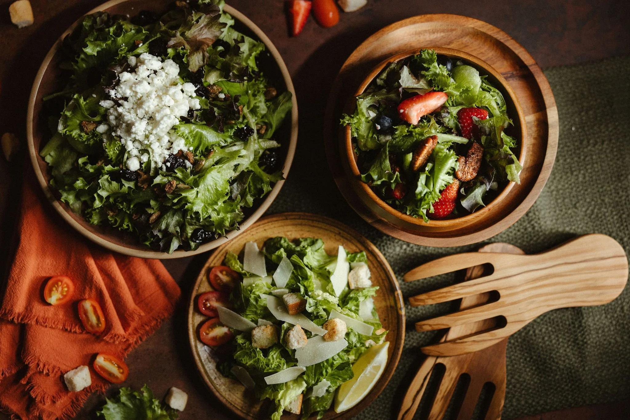 Several bowls of fresh salads with leafy greens, strawberries, blueberries, cherry tomatoes, croutons, cheese, and other vegetables on a dark wooden table, with a wooden salad serving platter and an orange cloth napkin nearby.