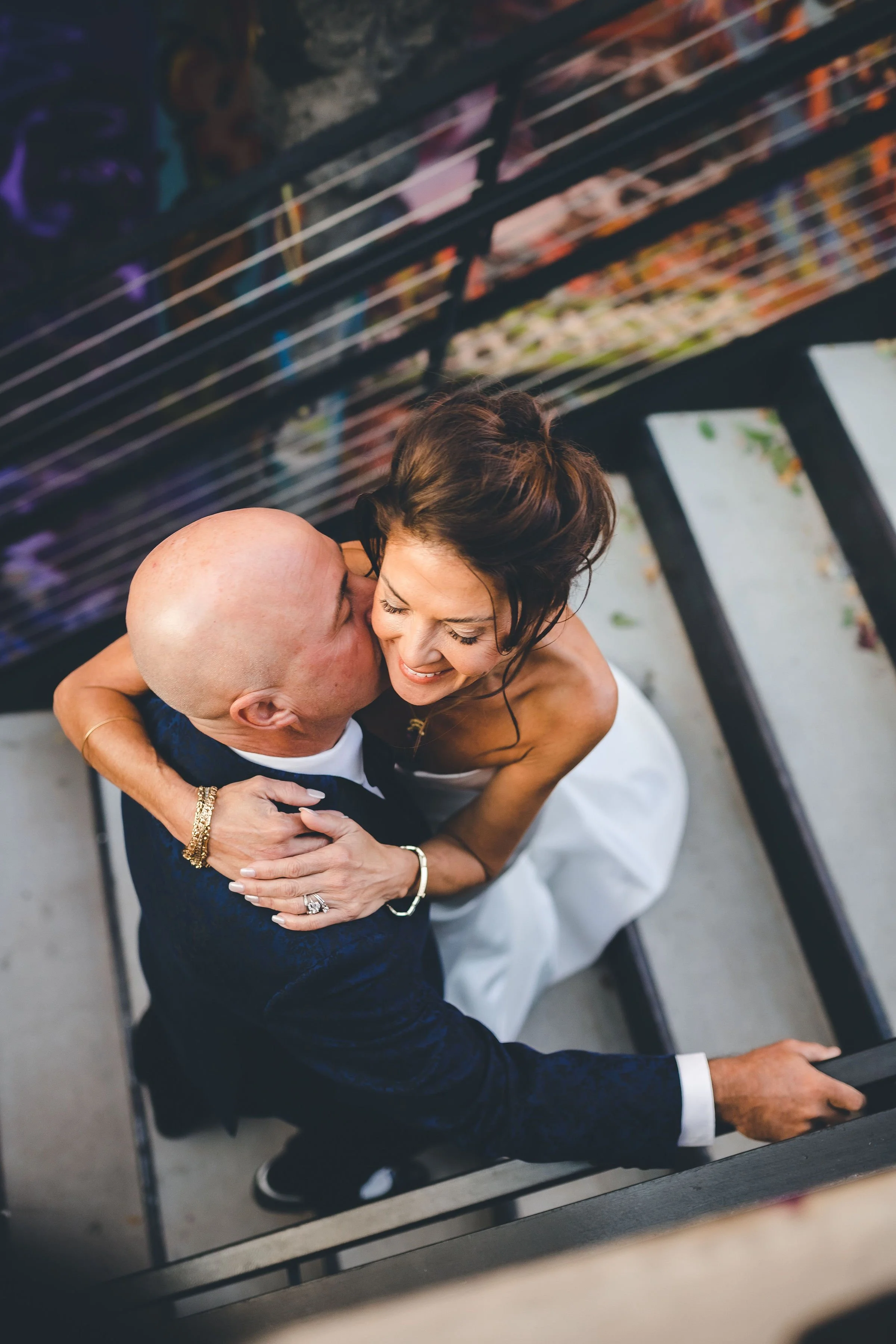 A couple sharing a kiss, taken from a mirror reflection. The woman has brown hair and is wearing a wedding dress, while the man is bald and dressed in a dark suit.