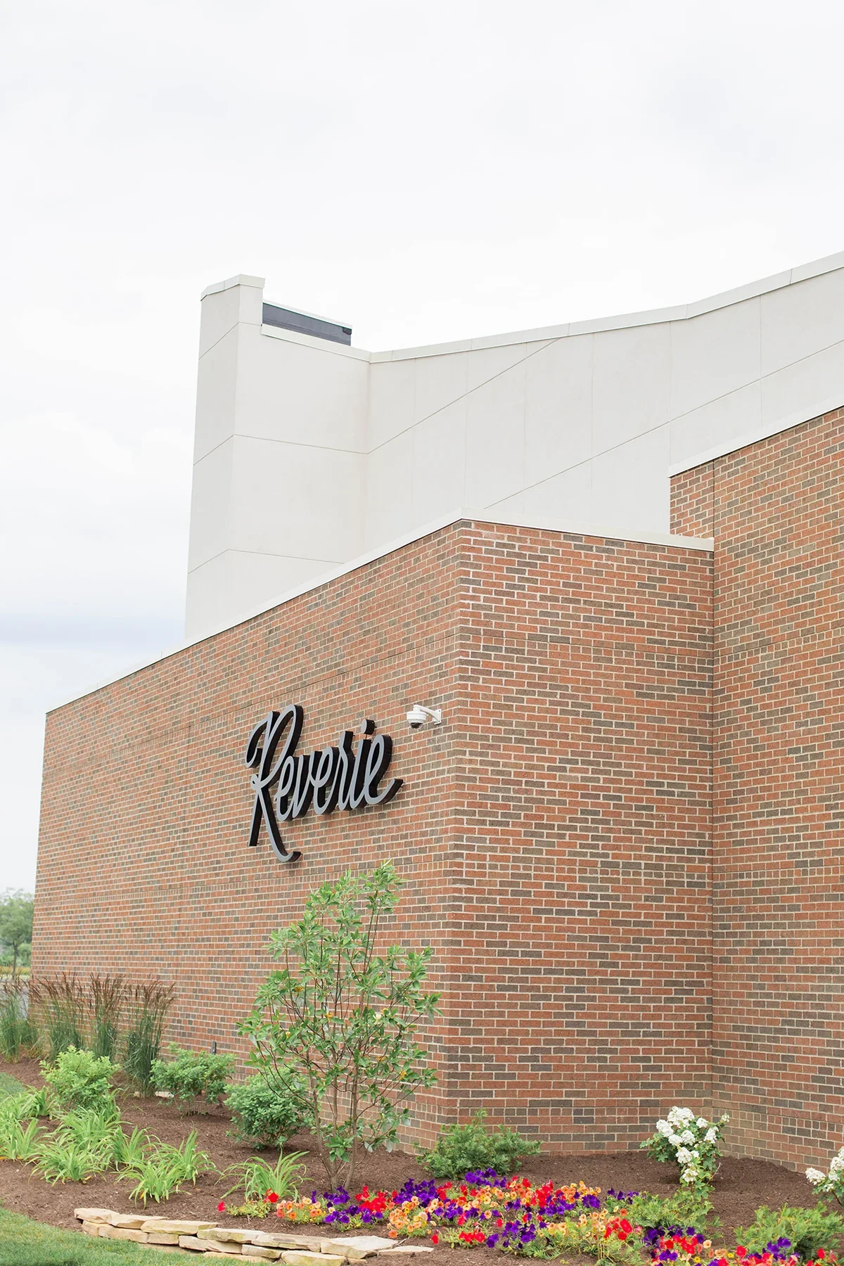 Exterior of a building with a brick facade, displaying a sign that says "Reverie" in black cursive letters. There are small plants and flowers in front of the building and a cloudy sky above.