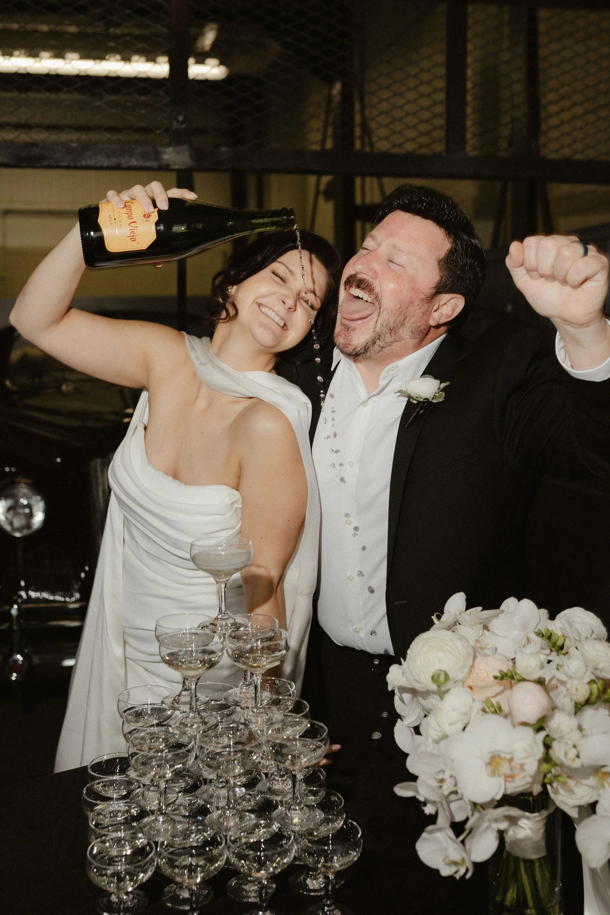 A bride and groom celebrating at a wedding reception, with the bride pouring champagne into a pyramid of glasses and the groom smiling and raising his fist in excitement. A large bouquet of white flowers is on the table in front of them.