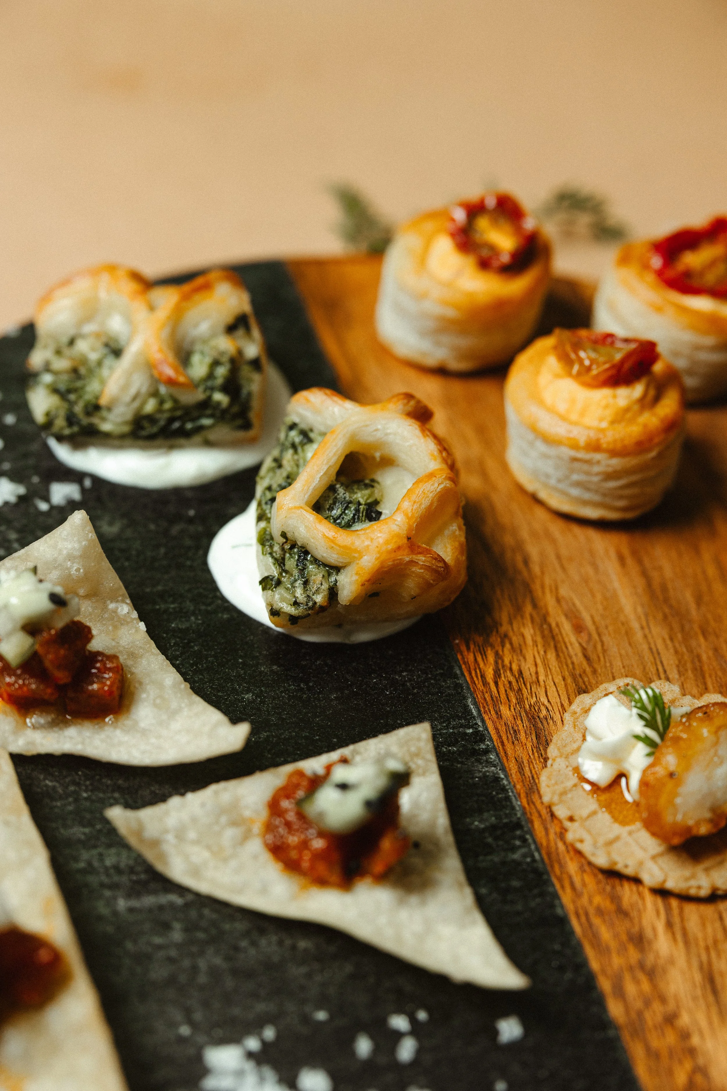 Close-up of assorted bite-sized appetizers, including puff pastry pinwheels with toppings, cracker with cream and garnish, and small pizza slices, arranged on a black slate and wooden serving platters.