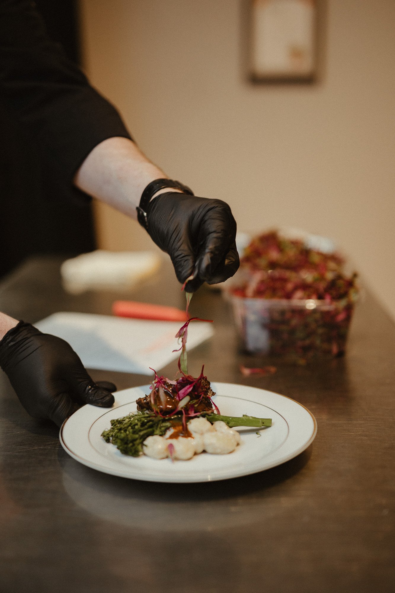 A person wearing black gloves and black clothing is plating a dish of gourmet food with tongs. The plate contains various food items garnished with microgreens, and there is a bowl of salad in the background.