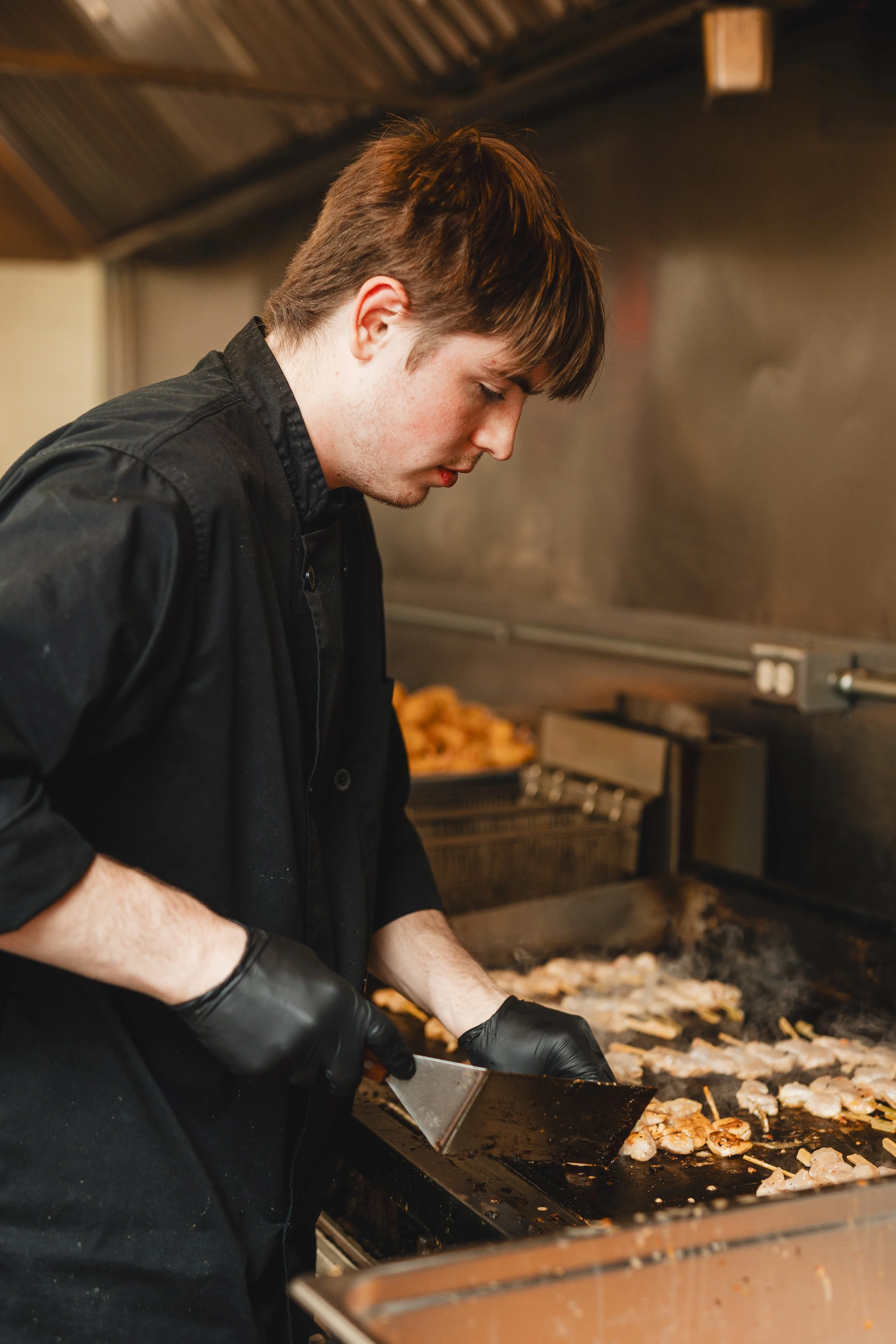 A young man with brown hair wearing a black shirt and black gloves is cooking on a flat-top griddle, preparing chicken and other ingredients in a kitchen.