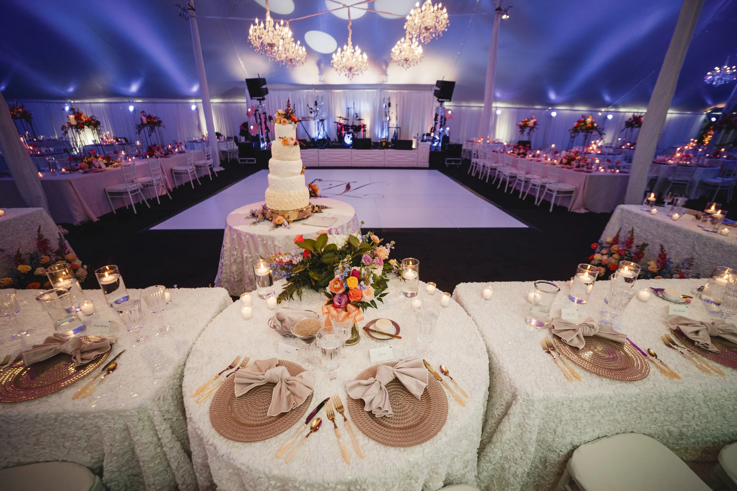 Elegant wedding reception inside a large white tent with chandeliers, decorated tables with floral centerpieces, candles, and a cake on a separate table in the background, ready for celebration.
