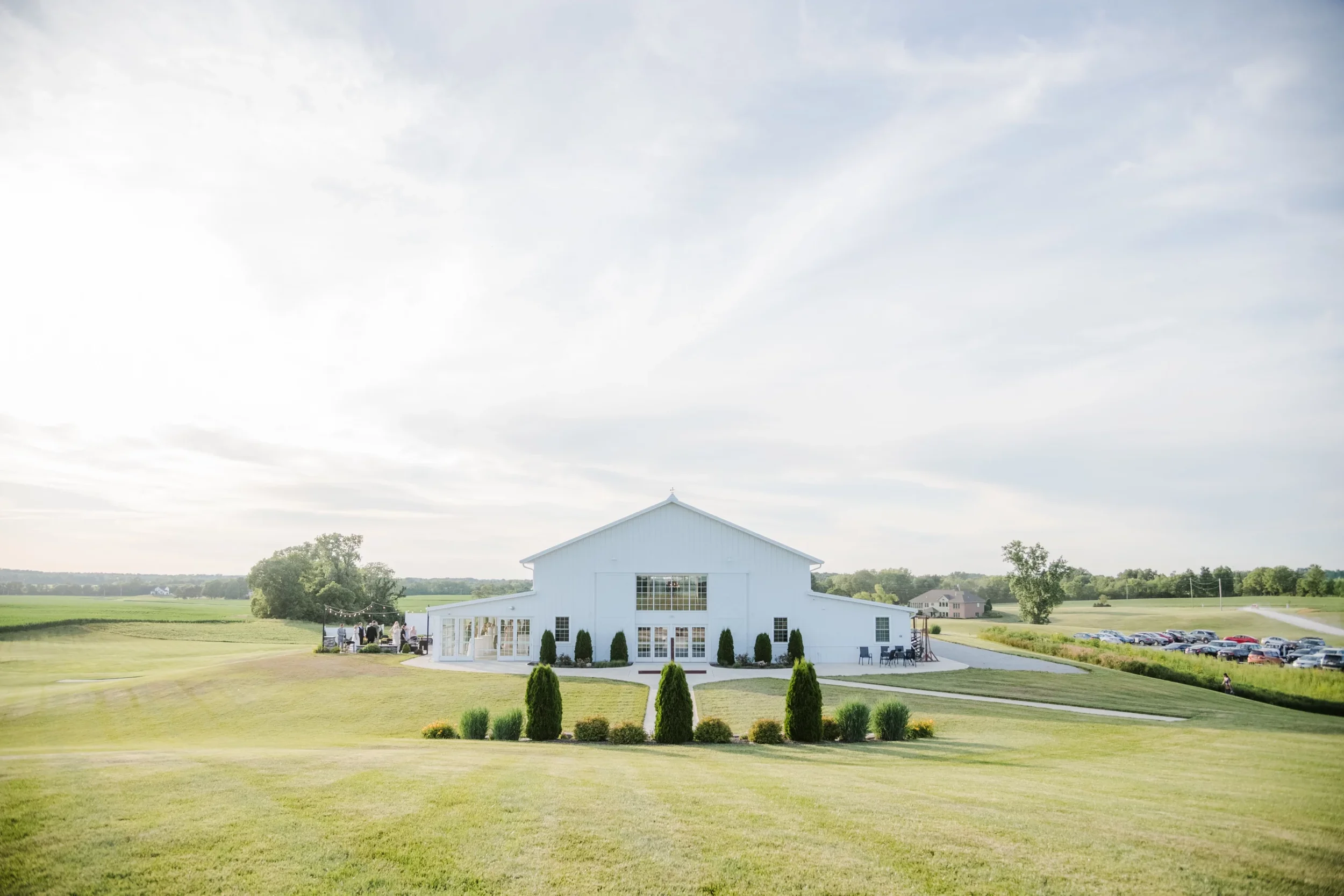 A large white barn in a green field with a group of people gathered outside, along with several cars parked nearby, and trees and other buildings in the background under a cloudy sky.