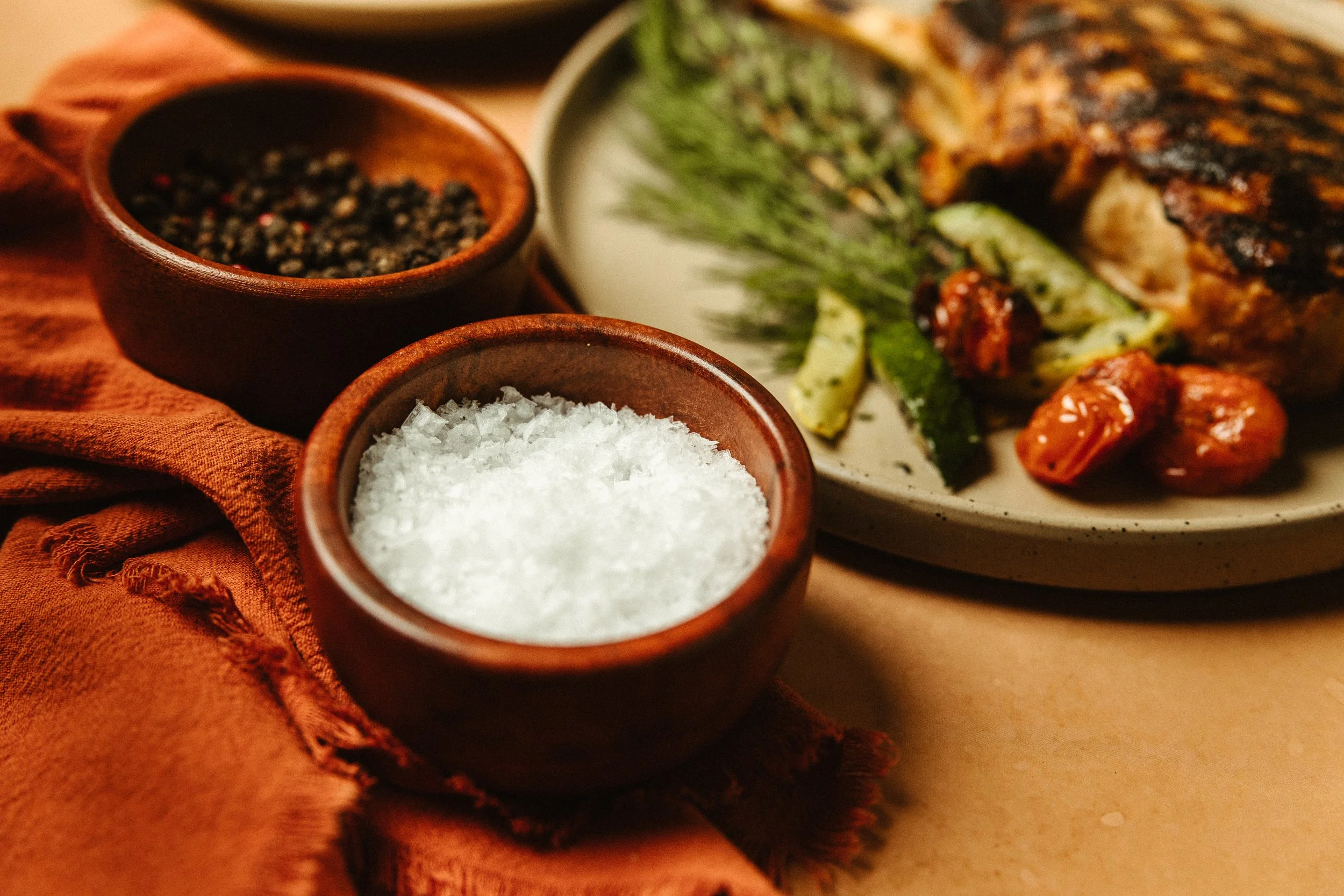 Close-up of two small wooden bowls filled with black pepper and sea salt, on a rustic orange cloth, with a salad plate in the background containing grilled vegetables and greens.