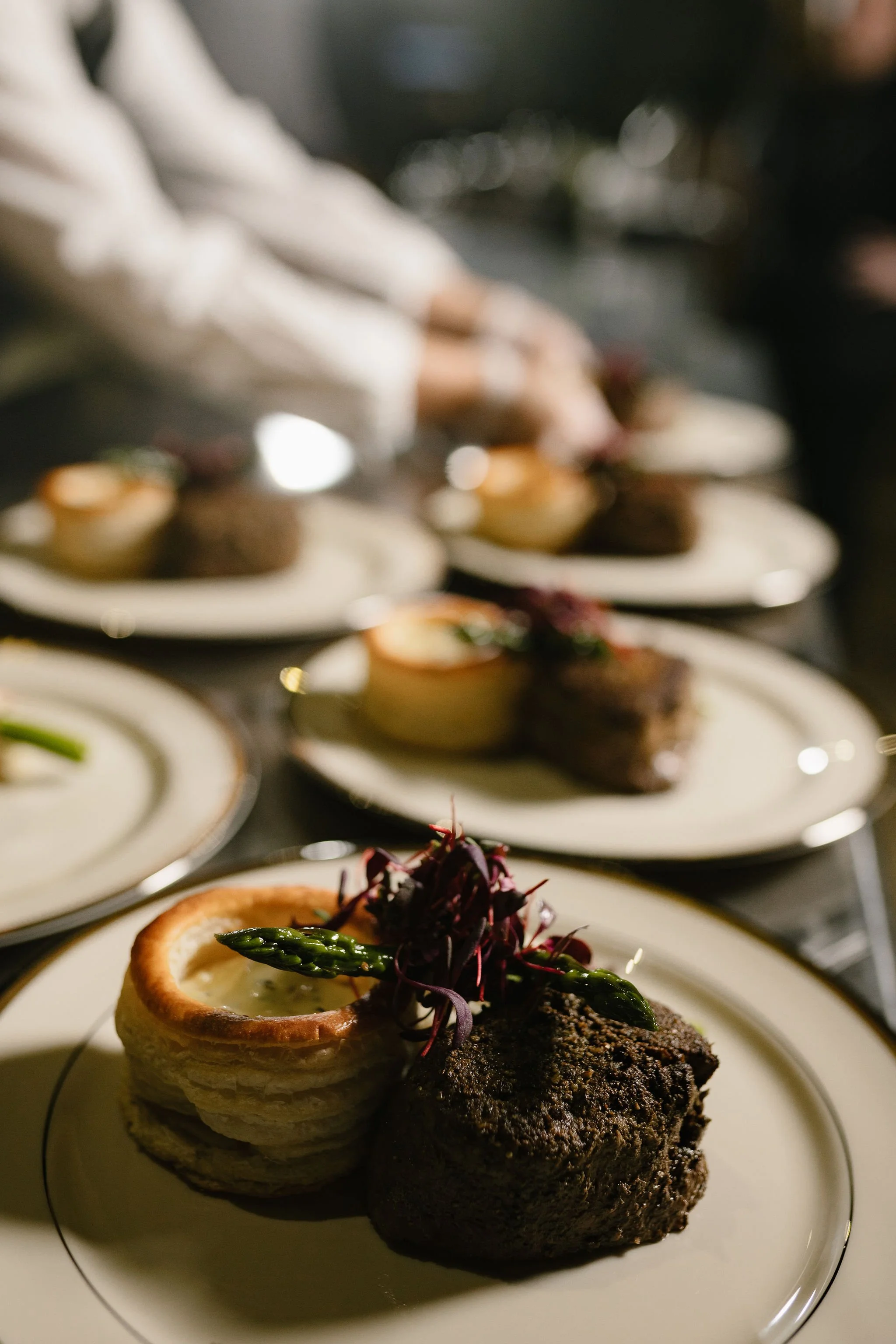 Close-up view of a plated dish featuring a steak topped with green asparagus tips, a small onion ring, and purple microgreens, with other similar dishes blurred in the background.