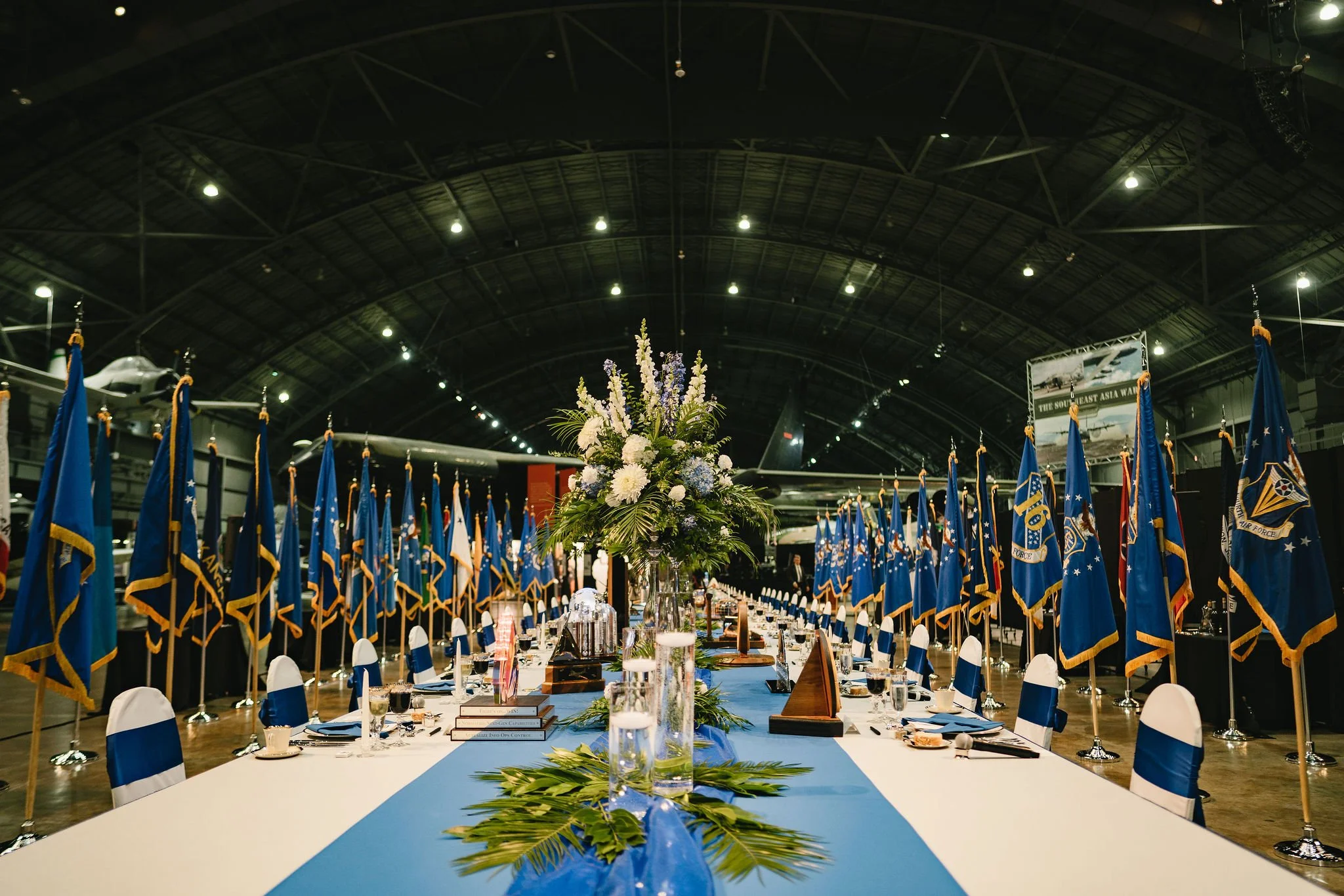 A banquet hall decorated for a formal event with a long table set with glassware and napkins, surrounded by blue flags, and a flower arrangement at the center of the table, in a large indoor venue with aircraft displayed overhead.