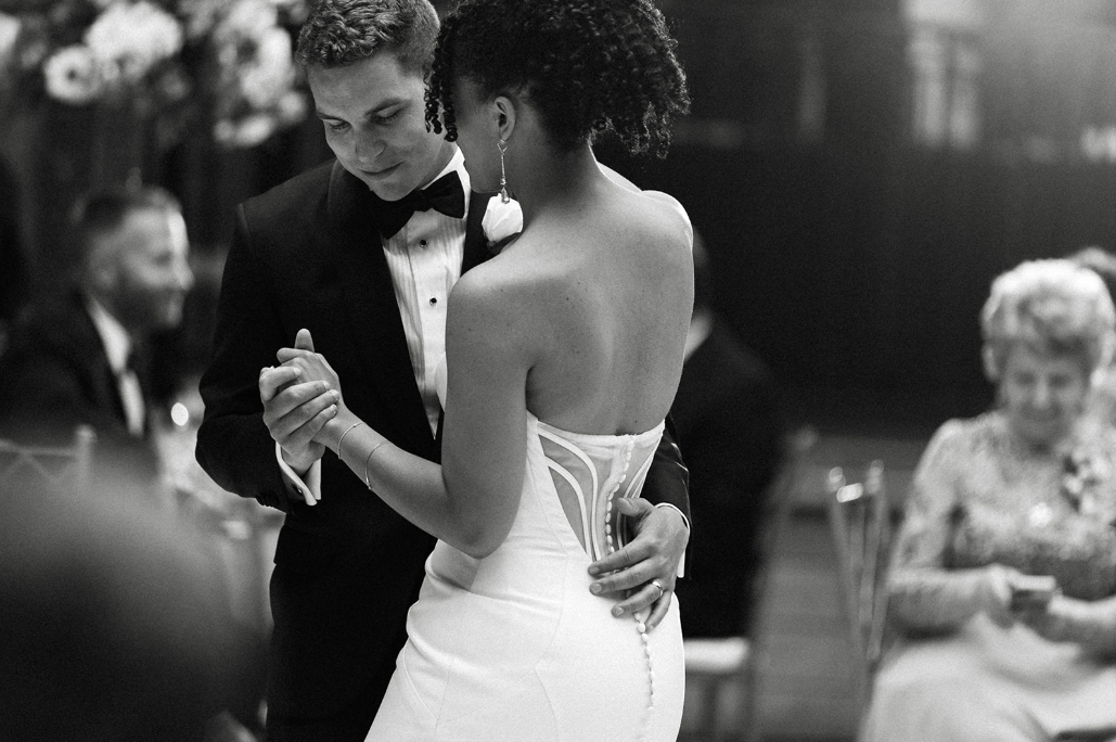 A black-and-white photo of a couple dancing closely at a formal event, with a woman in a strapless white gown and a man in a tuxedo, while other guests watch in the background.