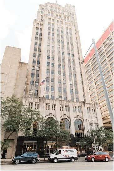 Tall skyscraper building with arched entrance, flags, and street parking in front.