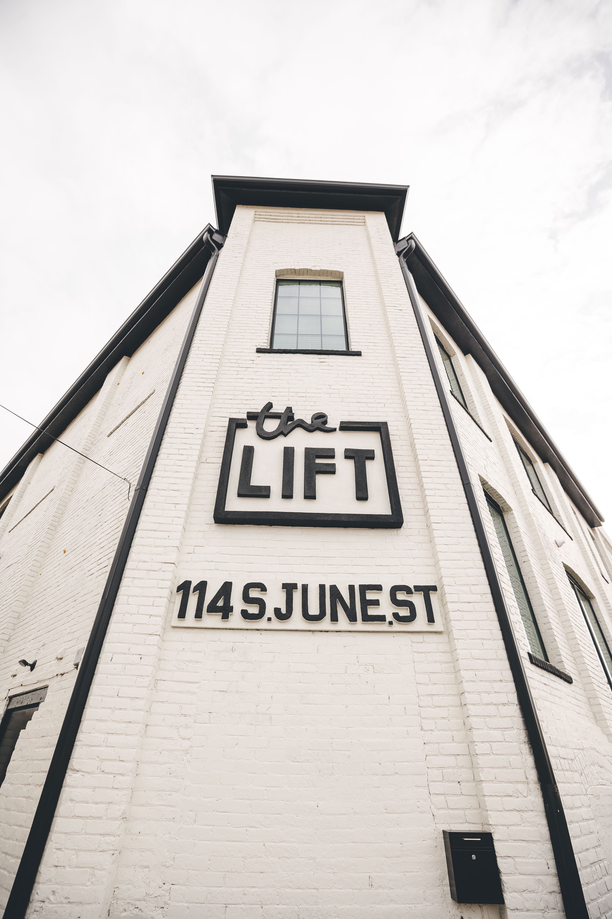 A white brick building with black signage reading 'the LIFT' and the address '114 S. JUNE ST', photographed from a low angle under an overcast sky.