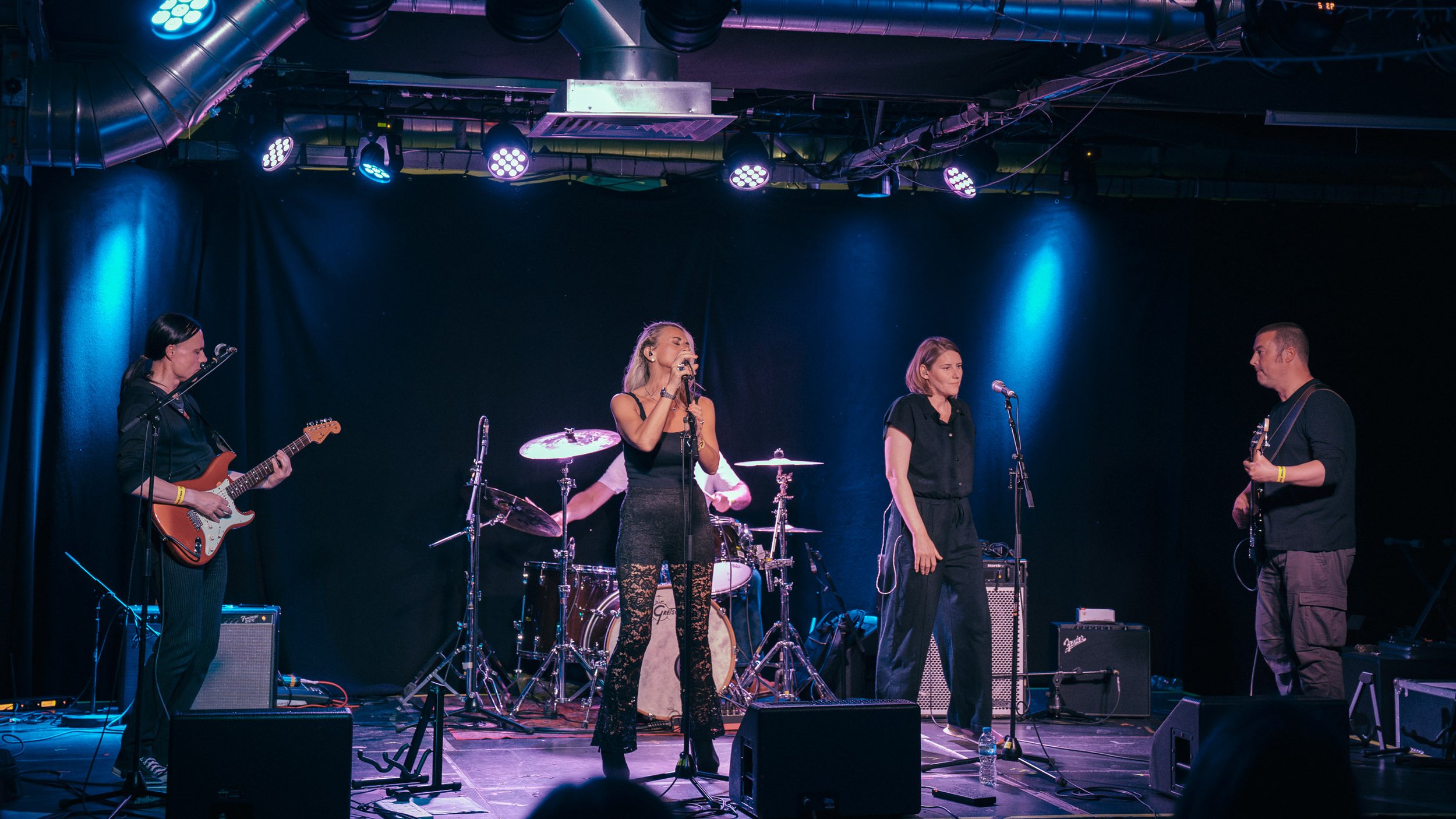 A band performing on stage with four members, including a female singer in the center, a guitarist on the left, a vocalist or speaker next to her, and a guitarist or bassist on the right, under stage lights.