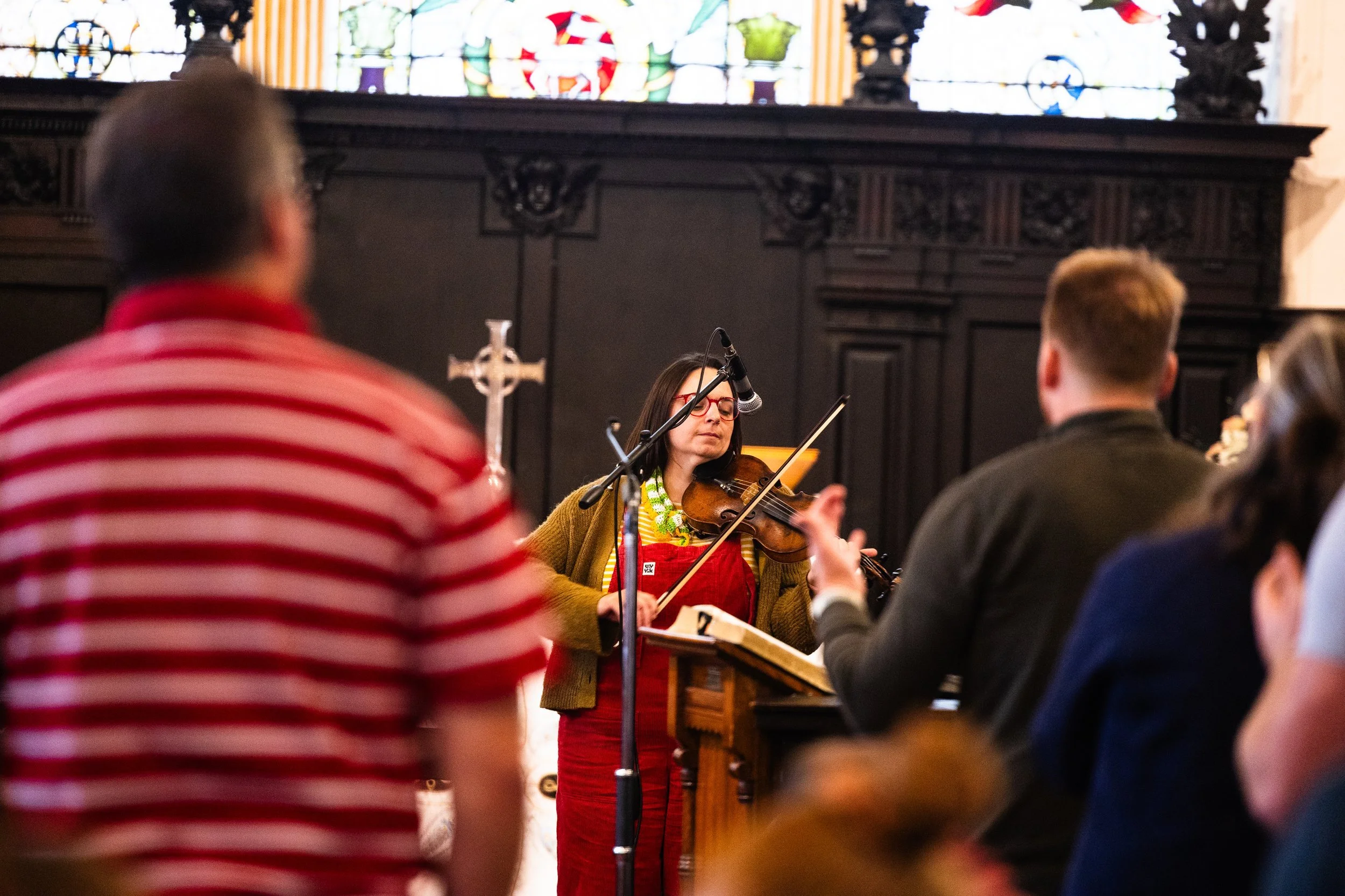 A woman playing the violin during a performance inside a church, with stained glass windows in the background, and several audience members in the foreground.