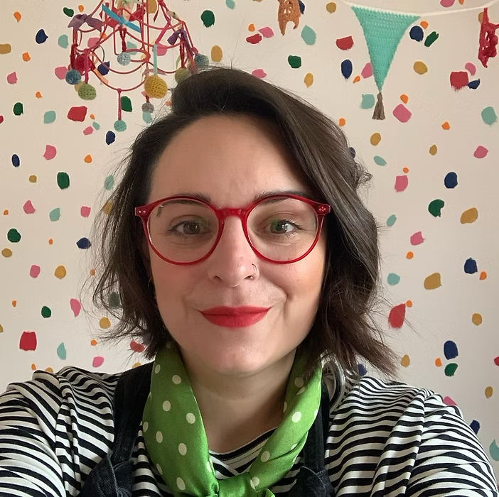 A woman taking a selfie in front of a white wall decorated with colorful confetti and bunting, wearing red glasses, a black and white striped shirt, and a green polka-dot scarf.