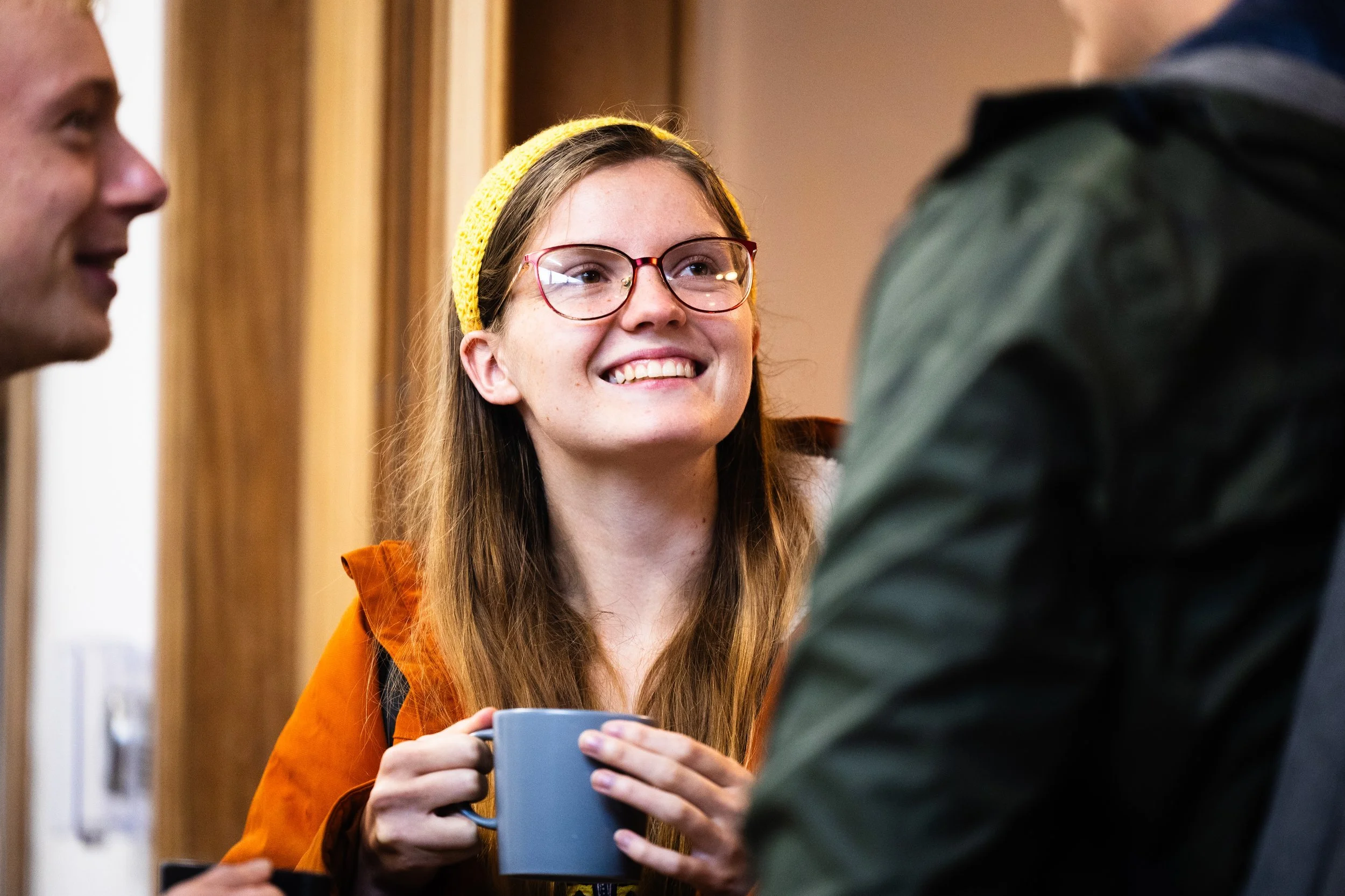 A young woman with glasses and a yellow knitted headband smiling while holding a blue coffee mug, engaging in conversation with two men, one partially visible, in an indoor setting.