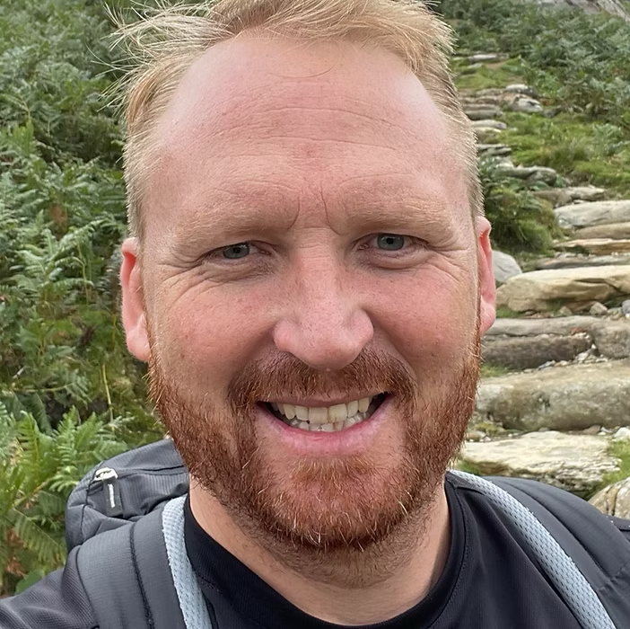 A smiling man with a beard and short blond hair outdoors with green plants and rocky terrain in the background.
