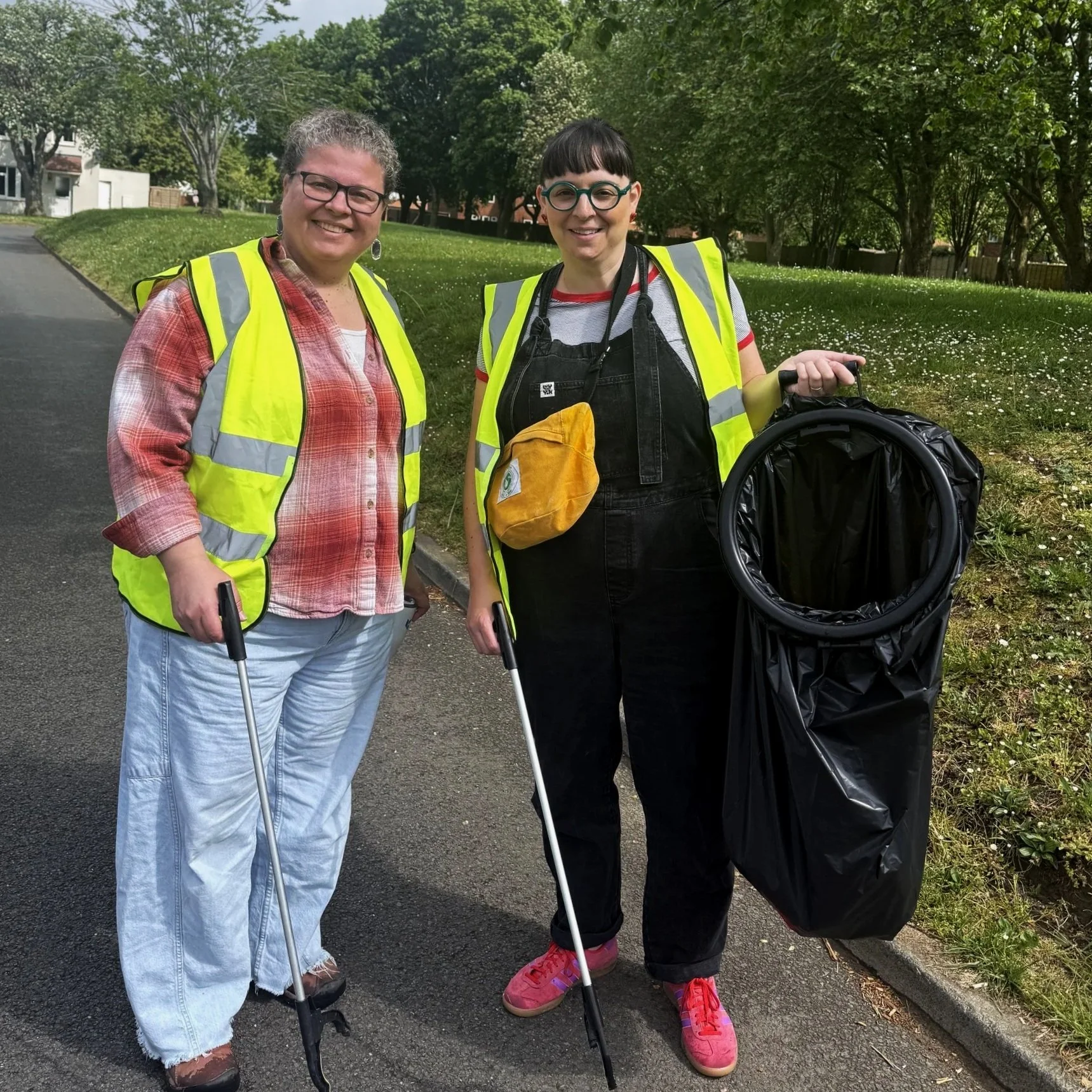 Two women wearing yellow reflective safety vests standing outside on a paved path, surrounded by green trees and grass, smiling and holding canes, one of whom has a large black bag hanging from her arm.