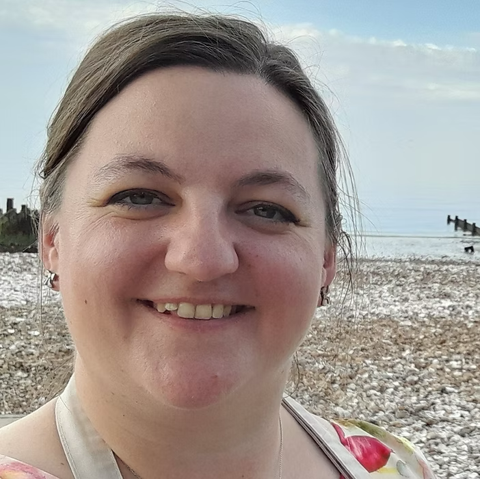 Close-up of a smiling woman with light brown hair, wearing a floral top, standing on a pebble beach with the ocean and sky in the background.