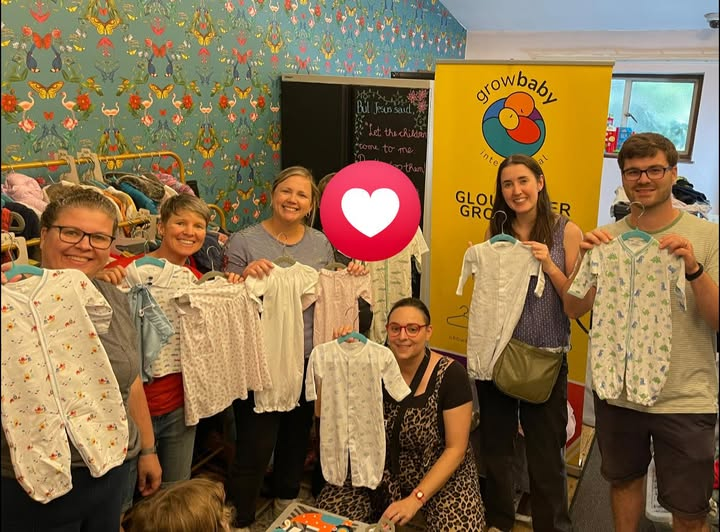 Group of five women and one man holding up baby clothes at an indoor event, with a colorful butterfly patterned wall and a yellow banner that reads 'growbaby' in the background.