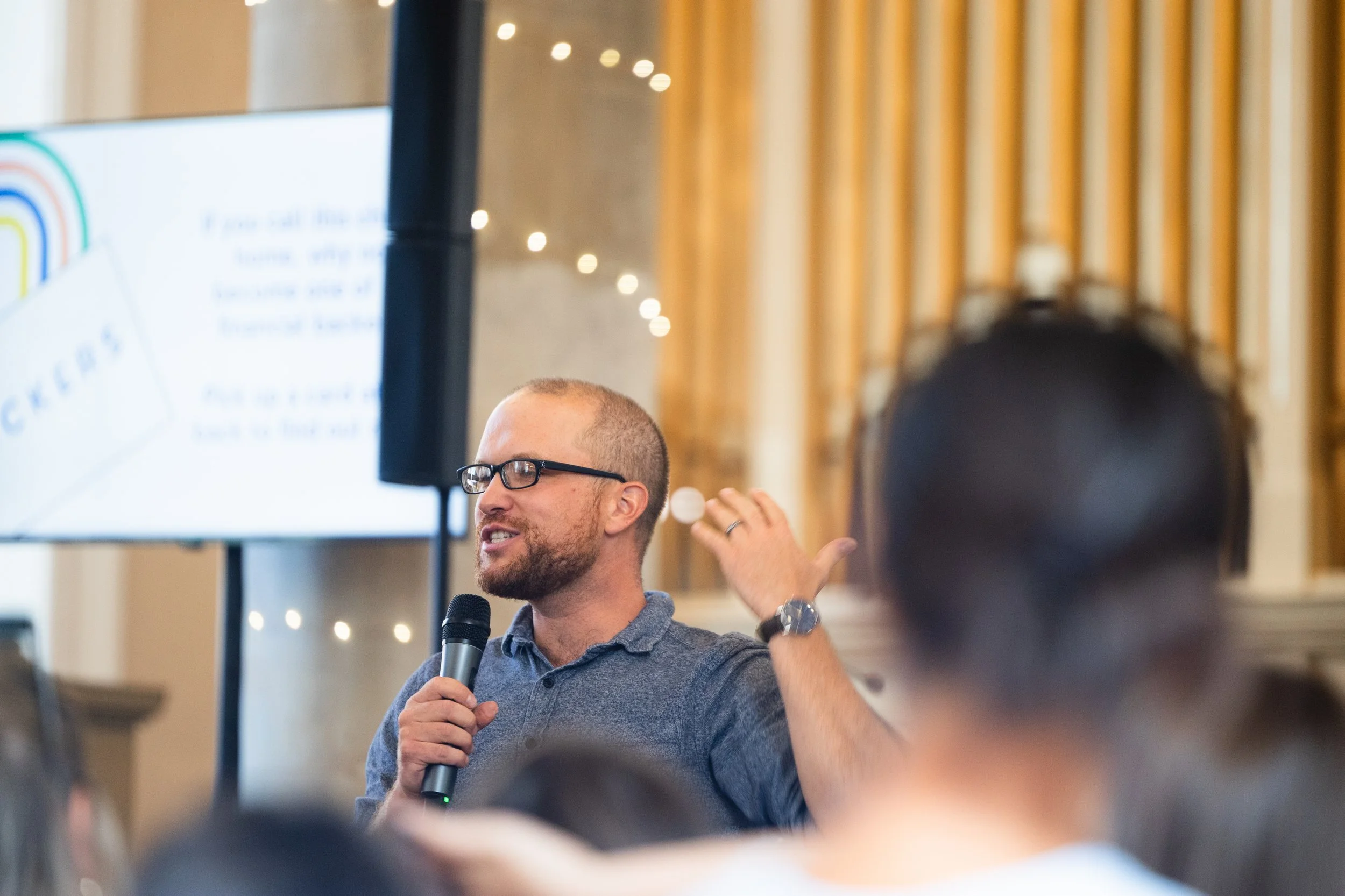 A man with glasses and a beard speaking into a microphone during a presentation, gesturing with his hand, with a blurred audience in the foreground and a large screen displaying text and colorful graphics behind him.