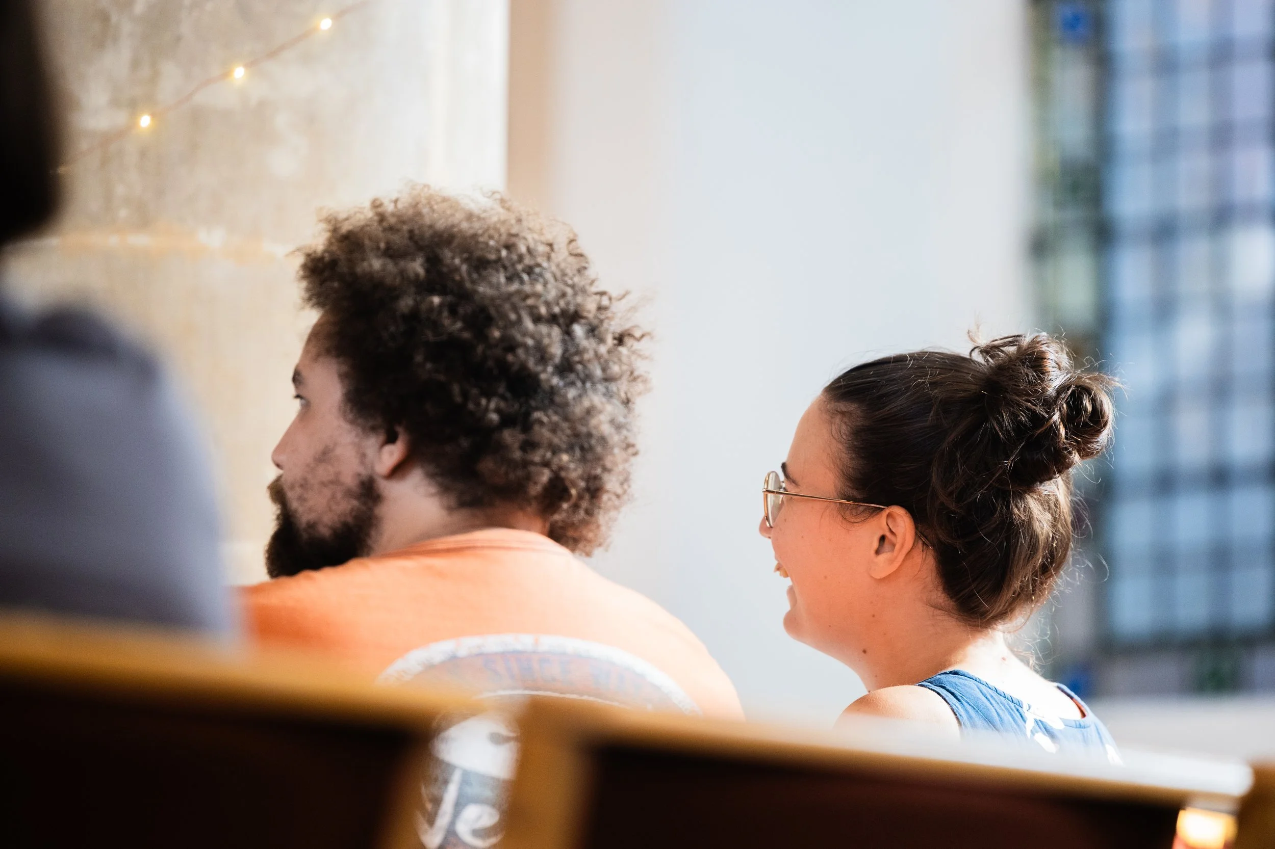 A group of diverse people sitting together, engaged in conversation or listening, with a blurred background of windows and lights.