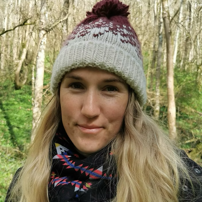 A woman with long blonde hair wearing a winter hat with a pom-pom, and a colorful scarf, standing outdoors in a wooded area.