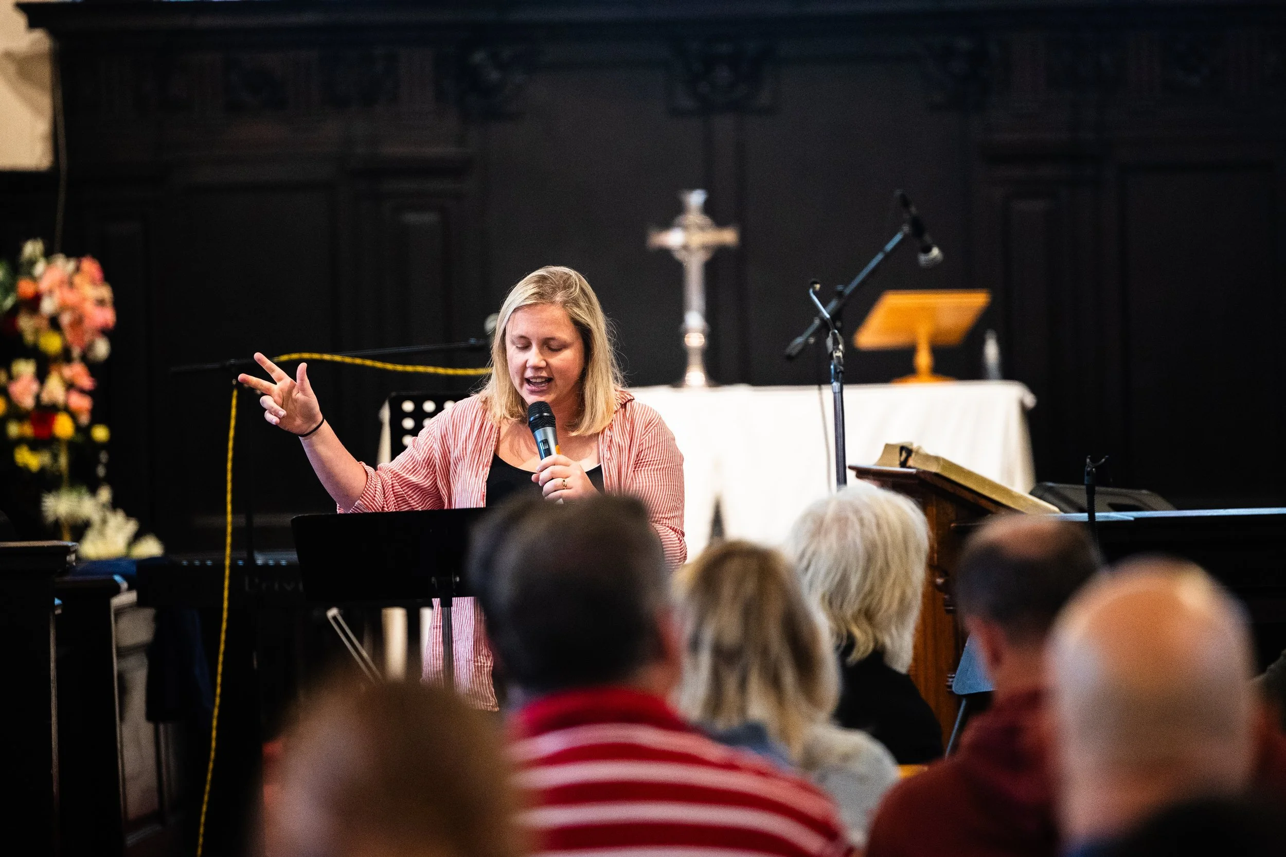 A woman in a striped shirt is speaking or singing into a microphone on stage during a performance or event. She has her eyes closed and one hand raised. There are microphones, music stands, and a table with a candlestick and cloth in the background, with an audience of children and adults seated in front.