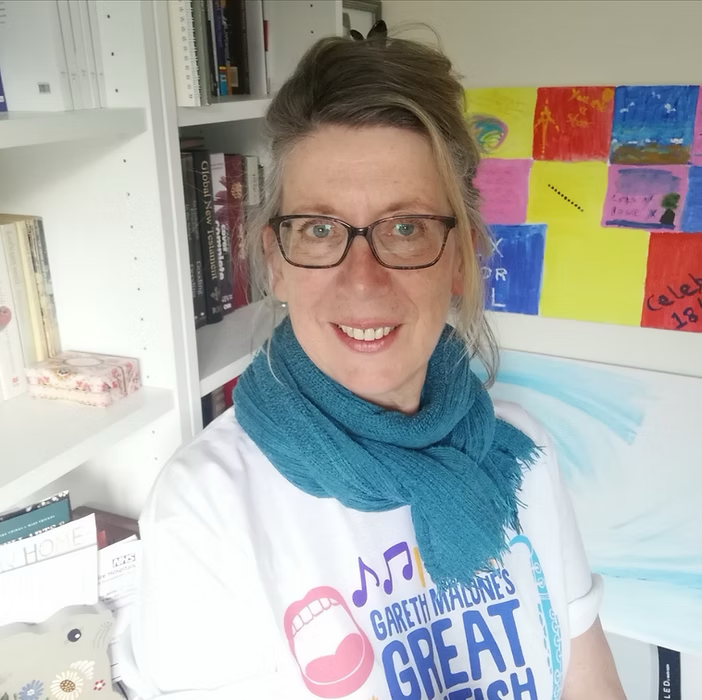 A smiling woman with glasses, wearing a white Gareth Malone's Great British Voice T-shirt and a blue scarf, standing in a room with white bookshelves, colorful artwork on the wall, and various books and items.
