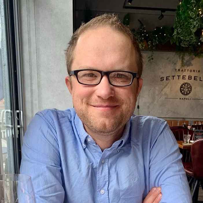 A man with glasses smiling, wearing a light blue button-up shirt, sitting at a restaurant.
