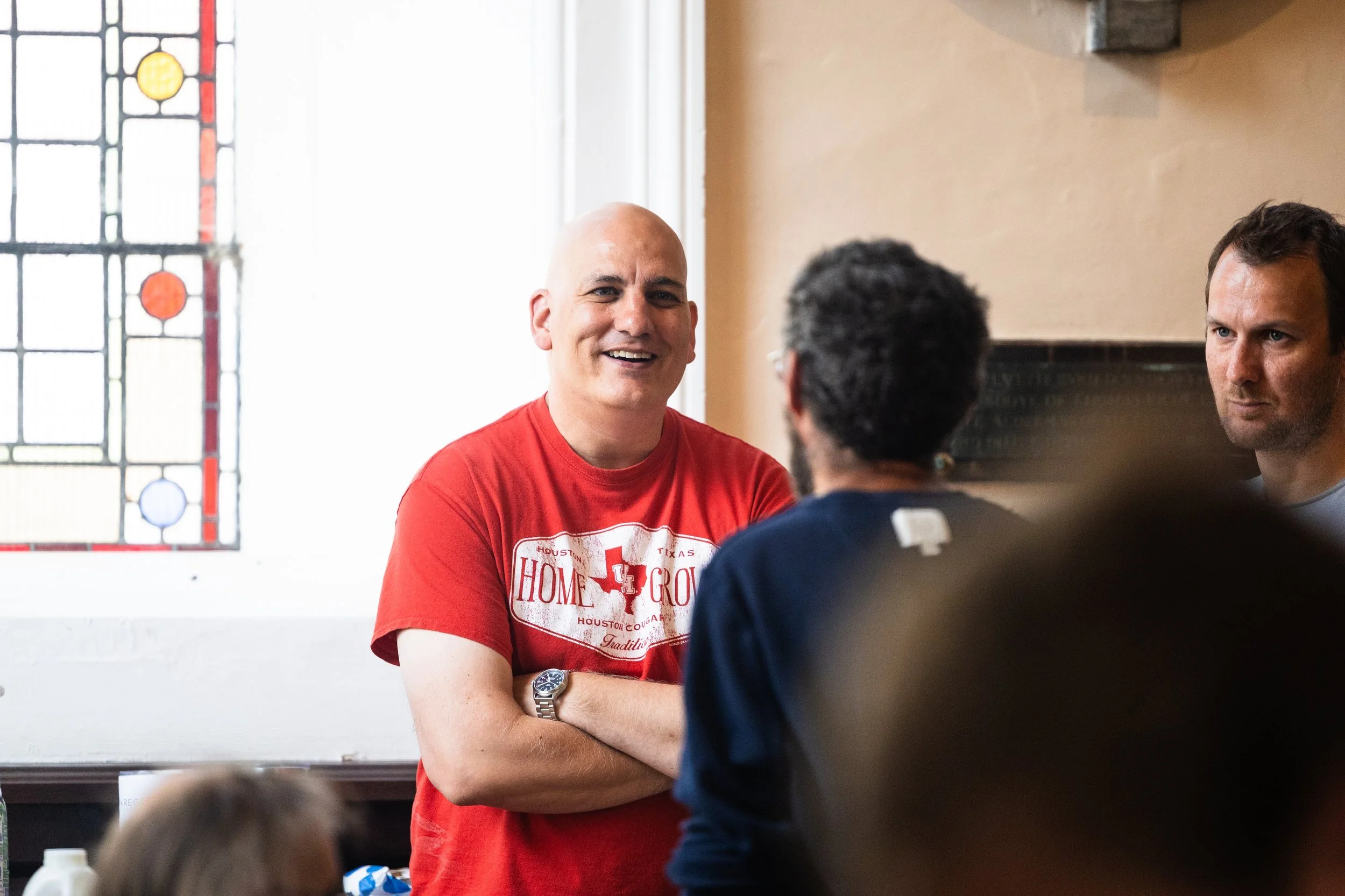 Three men are engaged in a conversation indoors. The man in the center is smiling, wearing a red t-shirt with text on it. The other two men are facing him, one with his back to the camera and the other with a serious expression. There is a stained glass window in the background.