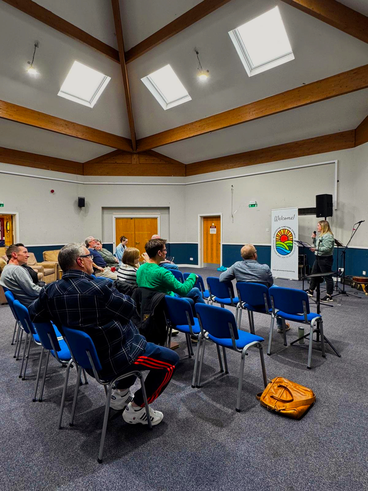 A woman giving a presentation to an audience in a conference room with wooden beams, skylights, and a sign that says "Welcome!"
